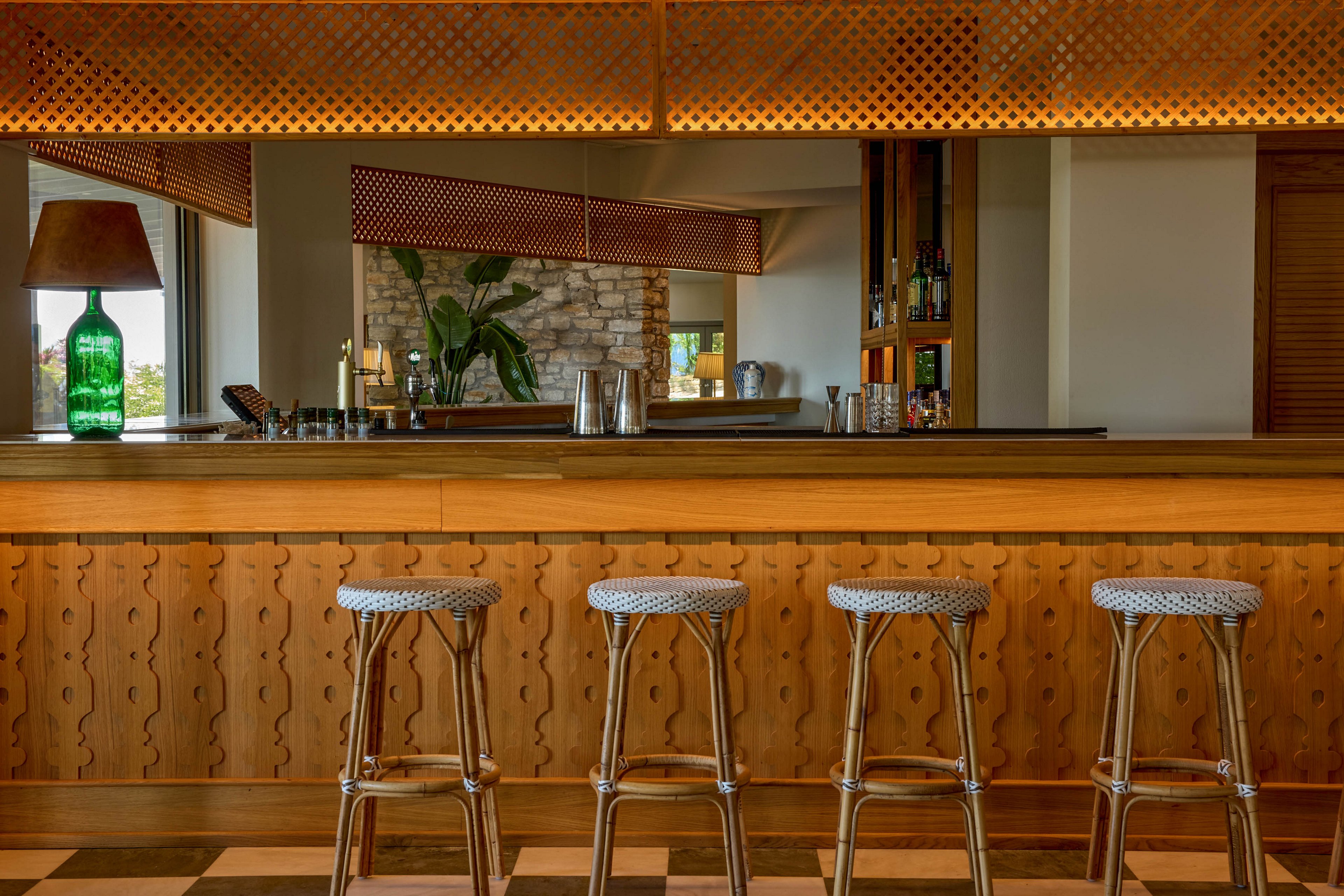 A cozy bar interior features a long wooden counter with four metal-framed bar stools neatly aligned. The setting includes warm lighting, a patterned wooden front, and a checkered floor that adds visual interest. In the background, a green glass bottle lamp, potted plant, and exposed brick wall contribute to the relaxed atmosphere. Shelves with bottles and bar tools are partially visible, suggesting a welcoming space for drinks and conversation.