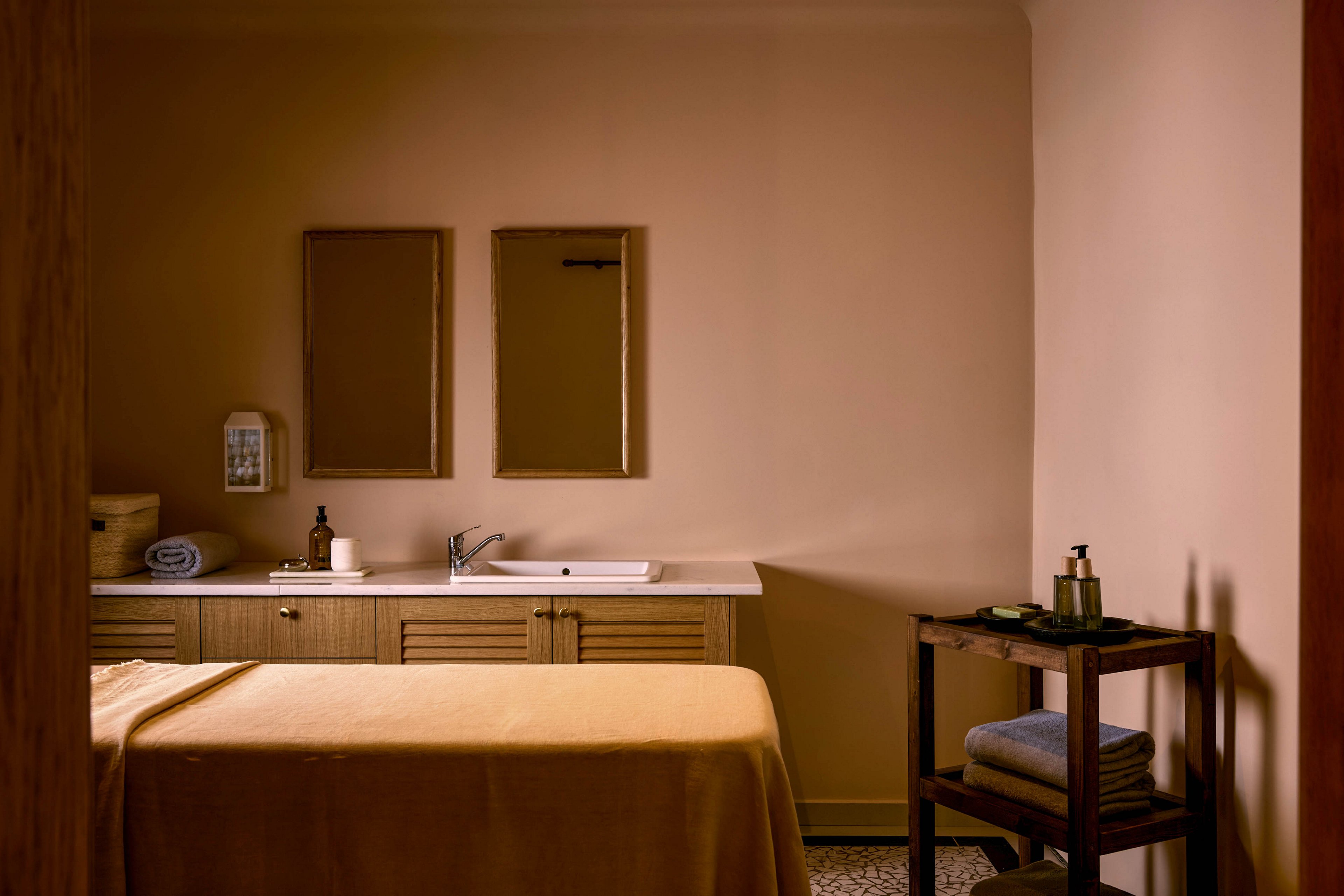 A cozy spa treatment room features a massage table covered in a warm beige sheet. At the back, a countertop with a sink, two rectangular mirrors, and neatly arranged toiletries creates a clean, organized look. A wooden cart holds folded towels and a soap dispenser, adding to the functional spa setup. Soft, warm lighting and neutral tones give the space a calm, relaxing atmosphere.