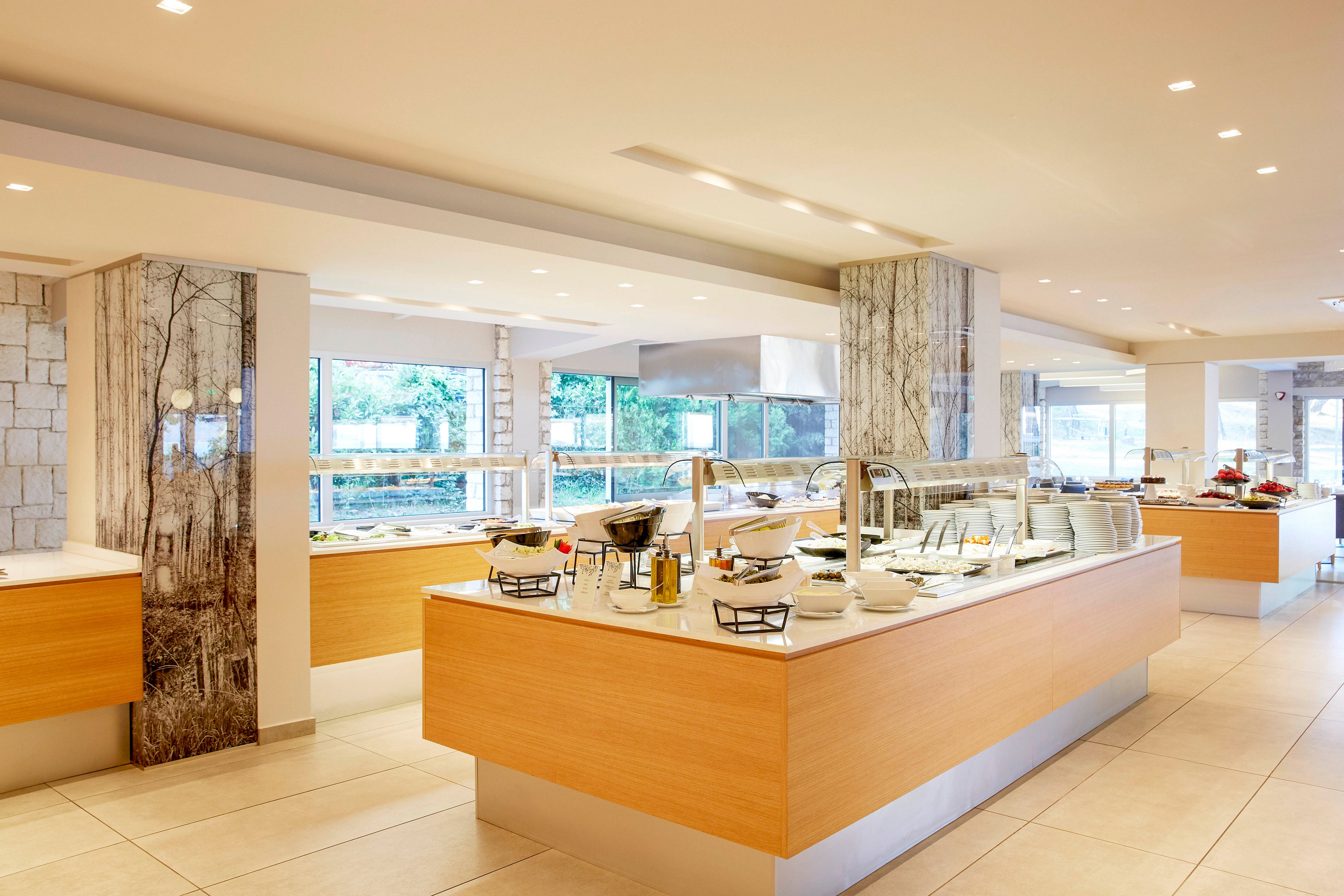 A contemporary self-service buffet area is shown in a bright, spacious dining hall. Light wood counters with glass sneeze guards display various dishes and serving utensils. Large windows bring in natural light and reveal greenery outside, while marble-clad columns add a refined touch. The overall scene is clean, organized, and ready for guests, emphasizing a modern hospitality environment.