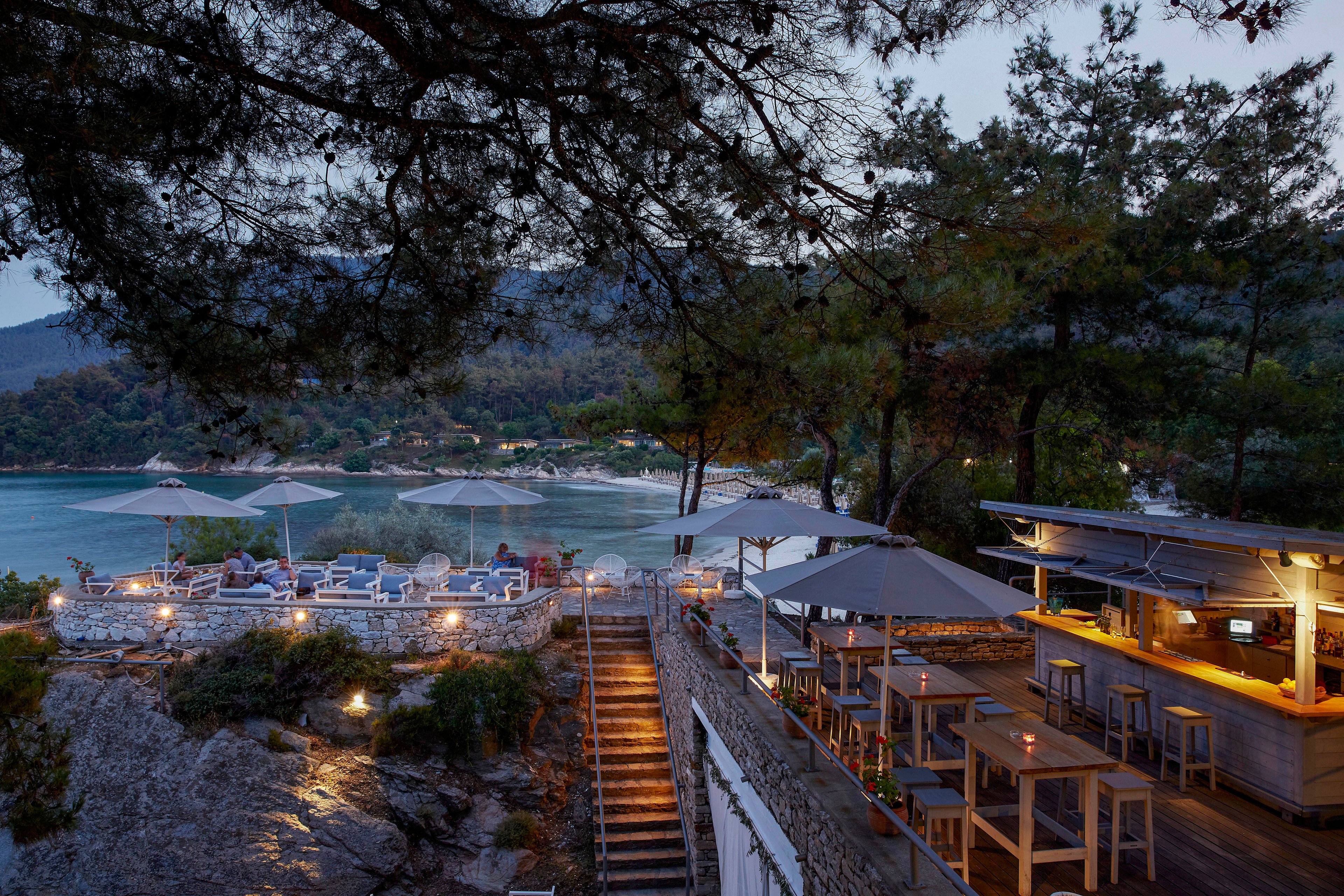 An open-air restaurant and bar sits on a rocky hillside overlooking a calm coastal bay at dusk. Wooden tables and stools line the terrace, while warm lights illuminate the bar and a staircase leading down the slope. In the background, guests relax under large umbrellas near the water, framed by pine trees and forested hills. The overall atmosphere is serene and inviting, with soft evening light reflecting off the sea.