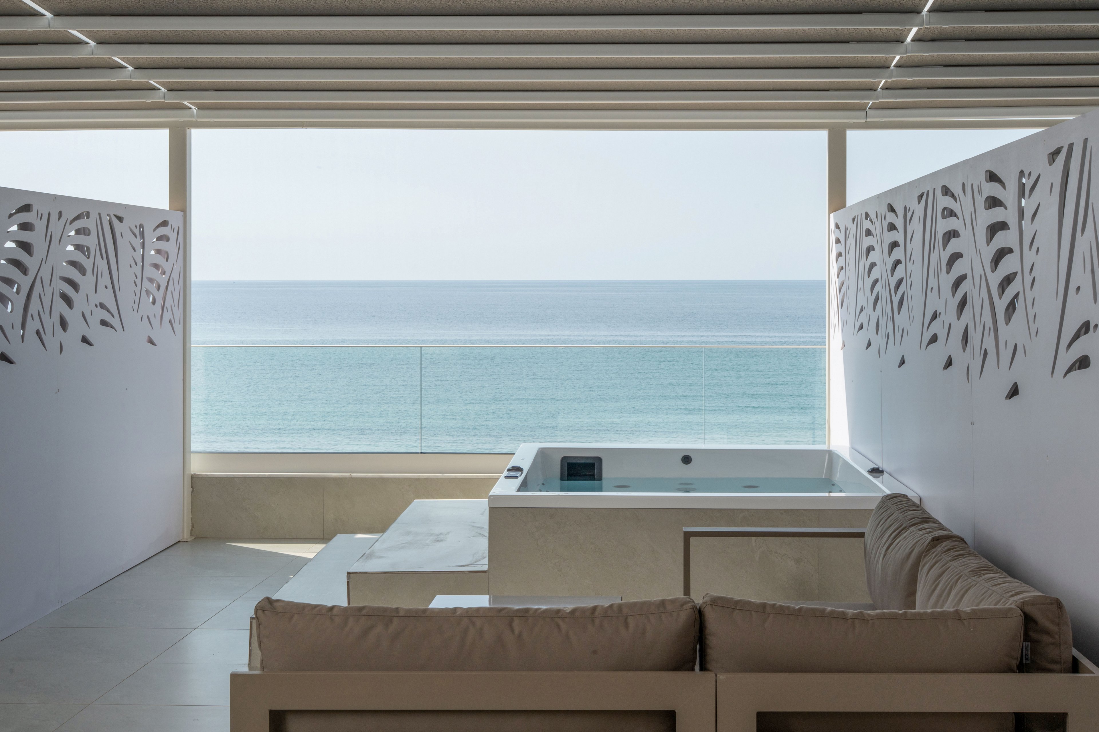 A contemporary terrace features a built-in jacuzzi facing a calm, expansive ocean view. Neutral-toned seating is arranged in the foreground, creating a comfortable lounge area. Decorative side panels with cut-out leaf patterns frame the scene, while horizontal slats above provide partial shade. The minimalist design and soft colors emphasize relaxation and a luxurious coastal atmosphere.