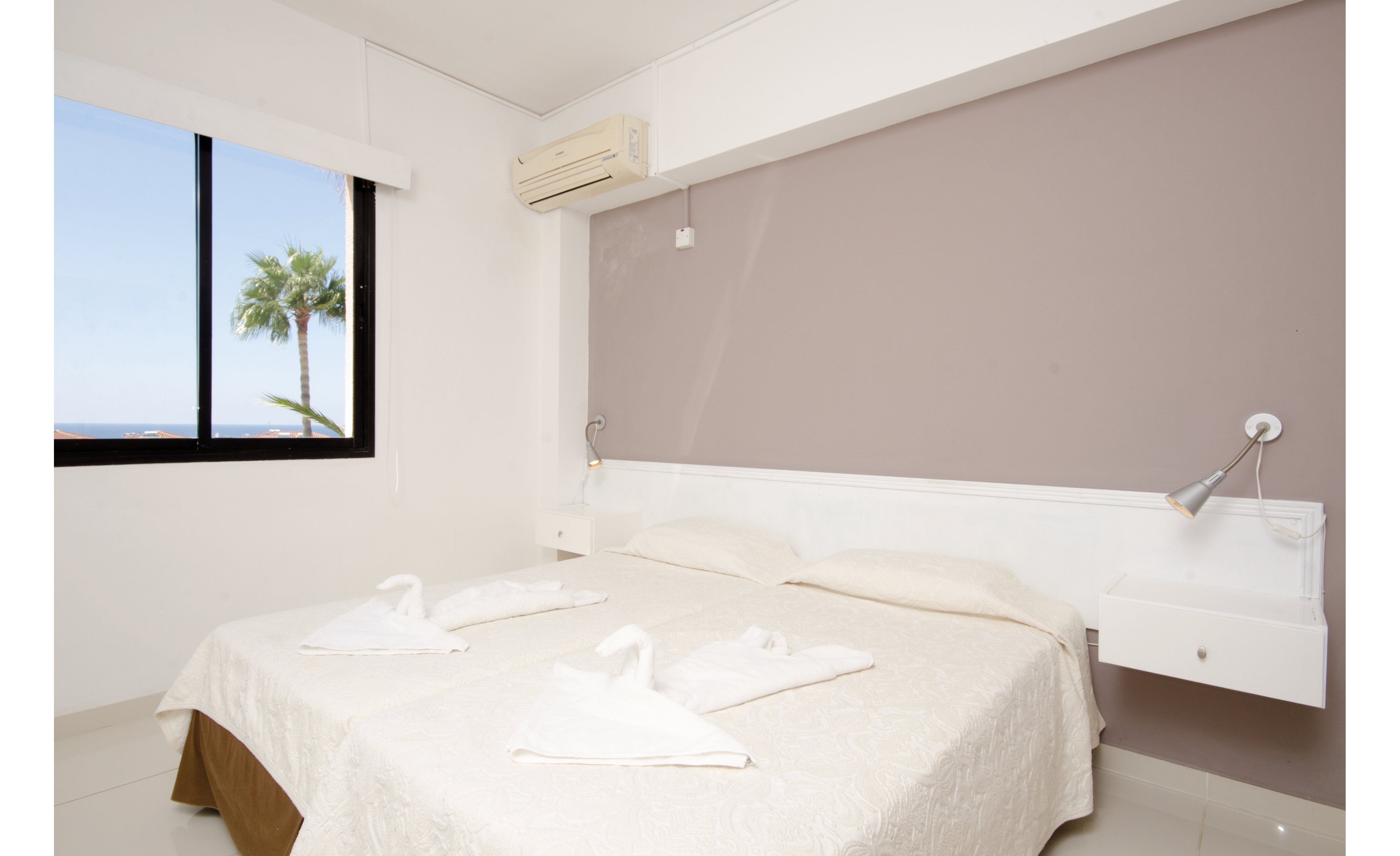 A clean and minimalistic hotel room featuring a double bed with white linens and towel swans. The room has a large window showcasing a palm tree and ocean view. Neutral tones dominate the decor, complemented by a wall-mounted air conditioner.