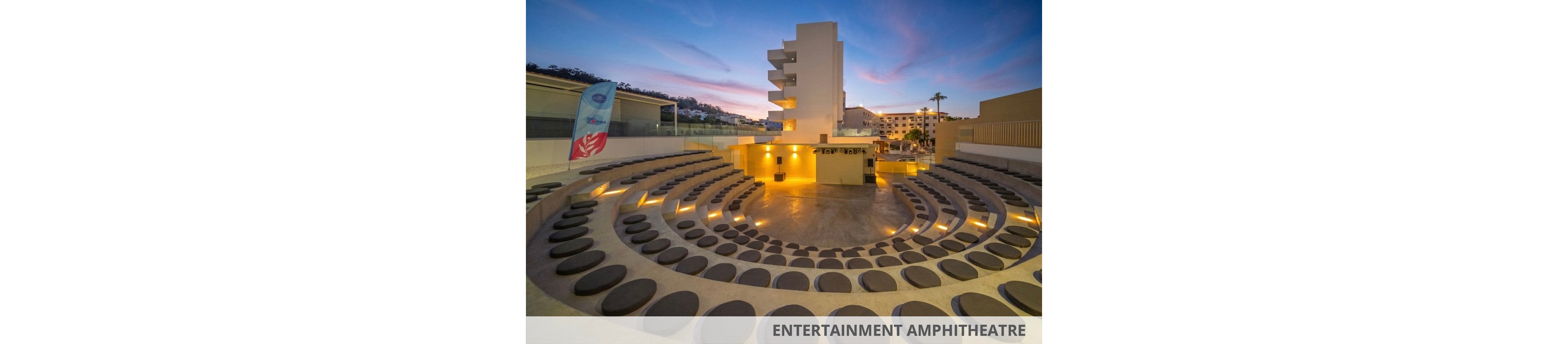 An empty circular outdoor amphitheater features tiered seating with round dark cushions arranged in concentric rows. Warm recessed lighting illuminates the steps and central stage area, creating a welcoming glow against the concrete surfaces. Modern buildings and a softly colored evening sky with streaked clouds form the backdrop. A vertical flag stands to the side, suggesting a resort or event venue setting.