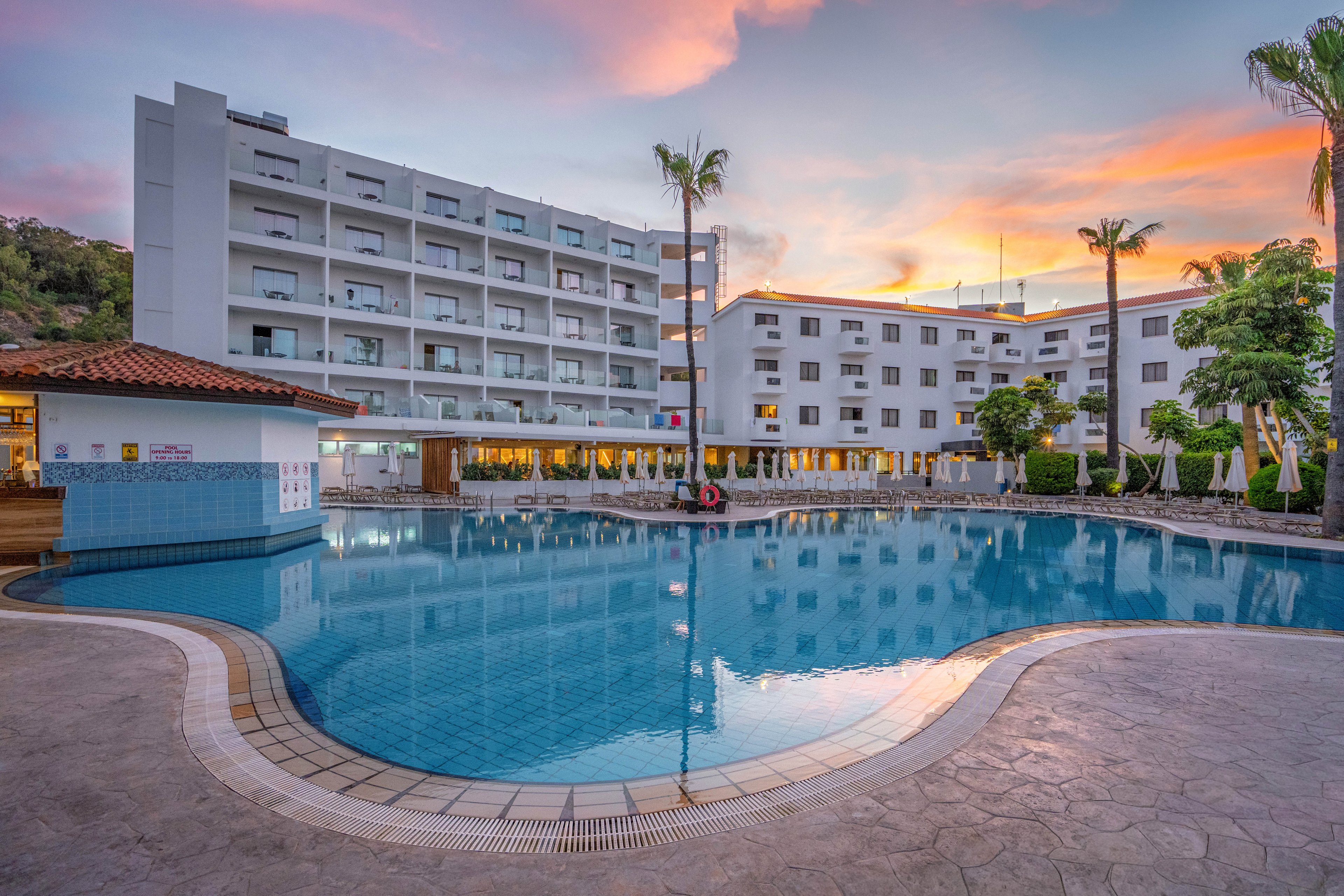 A modern multi-story resort hotel surrounds a large, calm swimming pool at dusk. The building features rows of balconies and warm interior lighting that reflects on the water. Palm trees and neatly trimmed greenery line the poolside, with sun loungers and umbrellas arranged along the edge. The sky is painted with soft pastel colors, enhancing the serene vacation atmosphere.