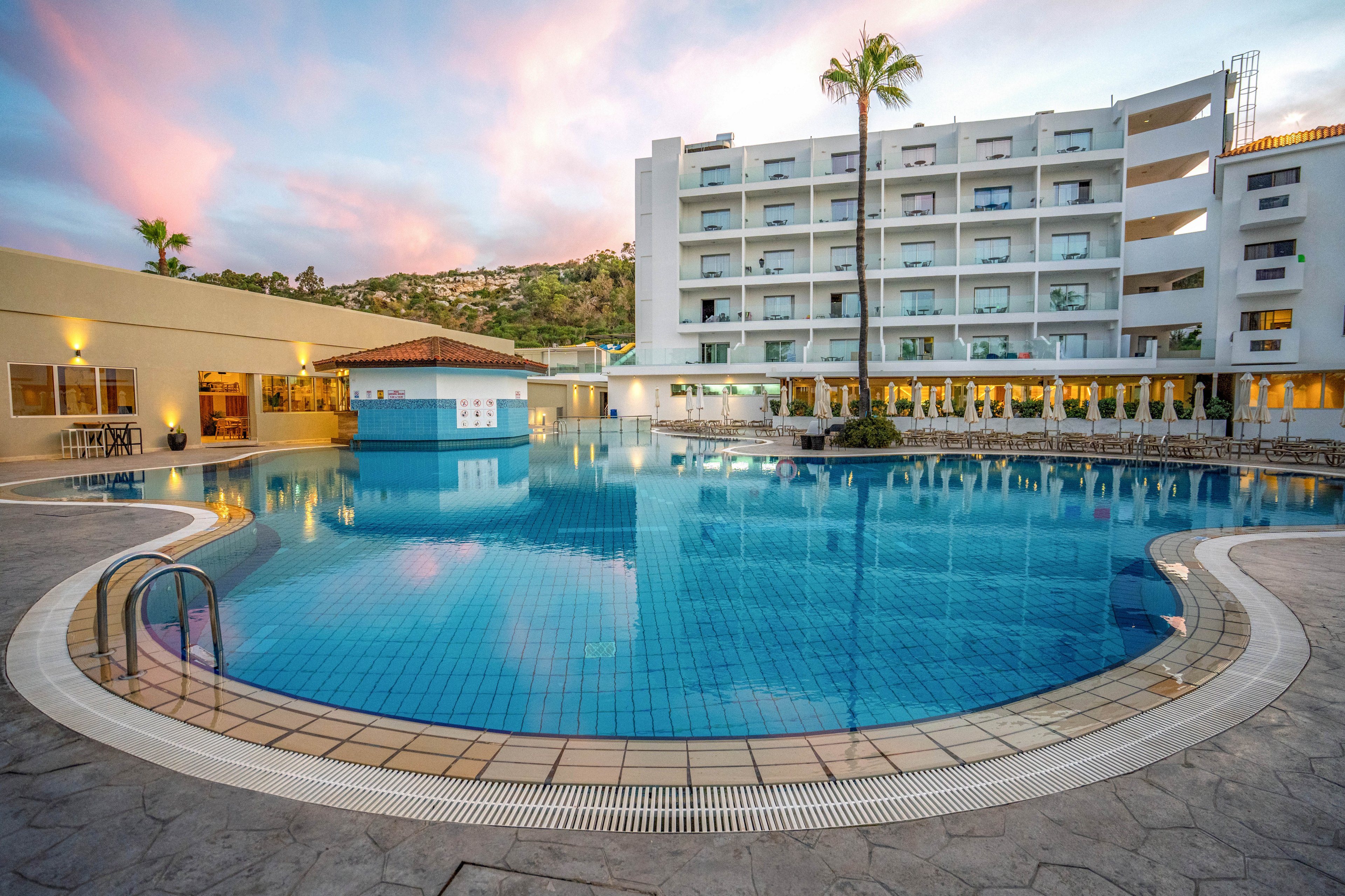 A contemporary multi-story hotel overlooks a large circular swimming pool at sunset. The clear blue water reflects the building, palm trees, and pastel sky. Pool ladders, tiled deck, and surrounding lounge areas suggest a relaxing vacation setting. Hillside scenery in the background adds to the resort atmosphere.