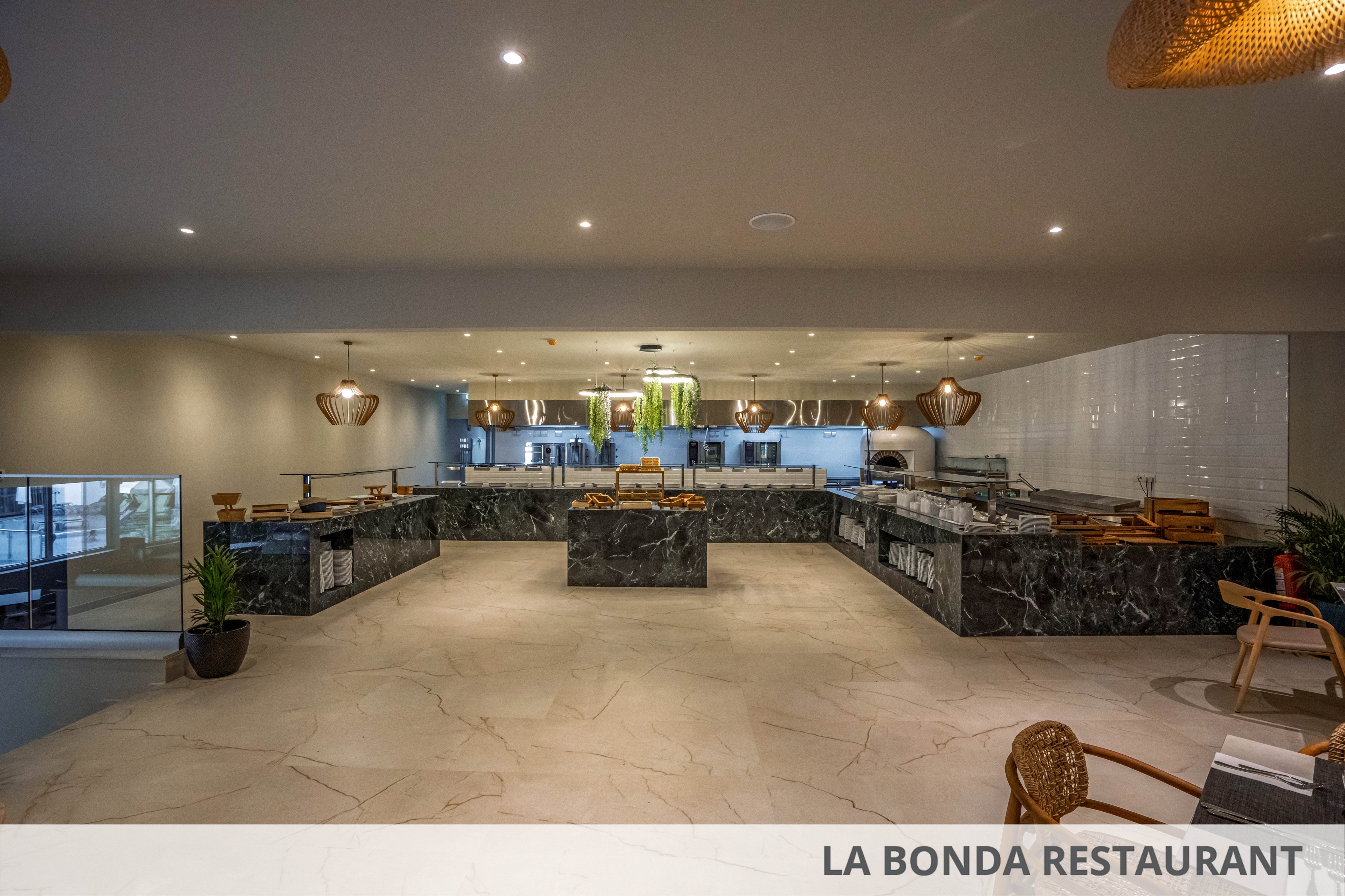A spacious, modern buffet restaurant features long black marble counters arranged in a U-shape. The room is well lit with recessed ceiling lights and warm hanging lamps. Food stations and serving dishes are arranged neatly along the counters, with plants adding a touch of greenery. Light stone flooring and neutral walls create a clean, contemporary dining atmosphere.