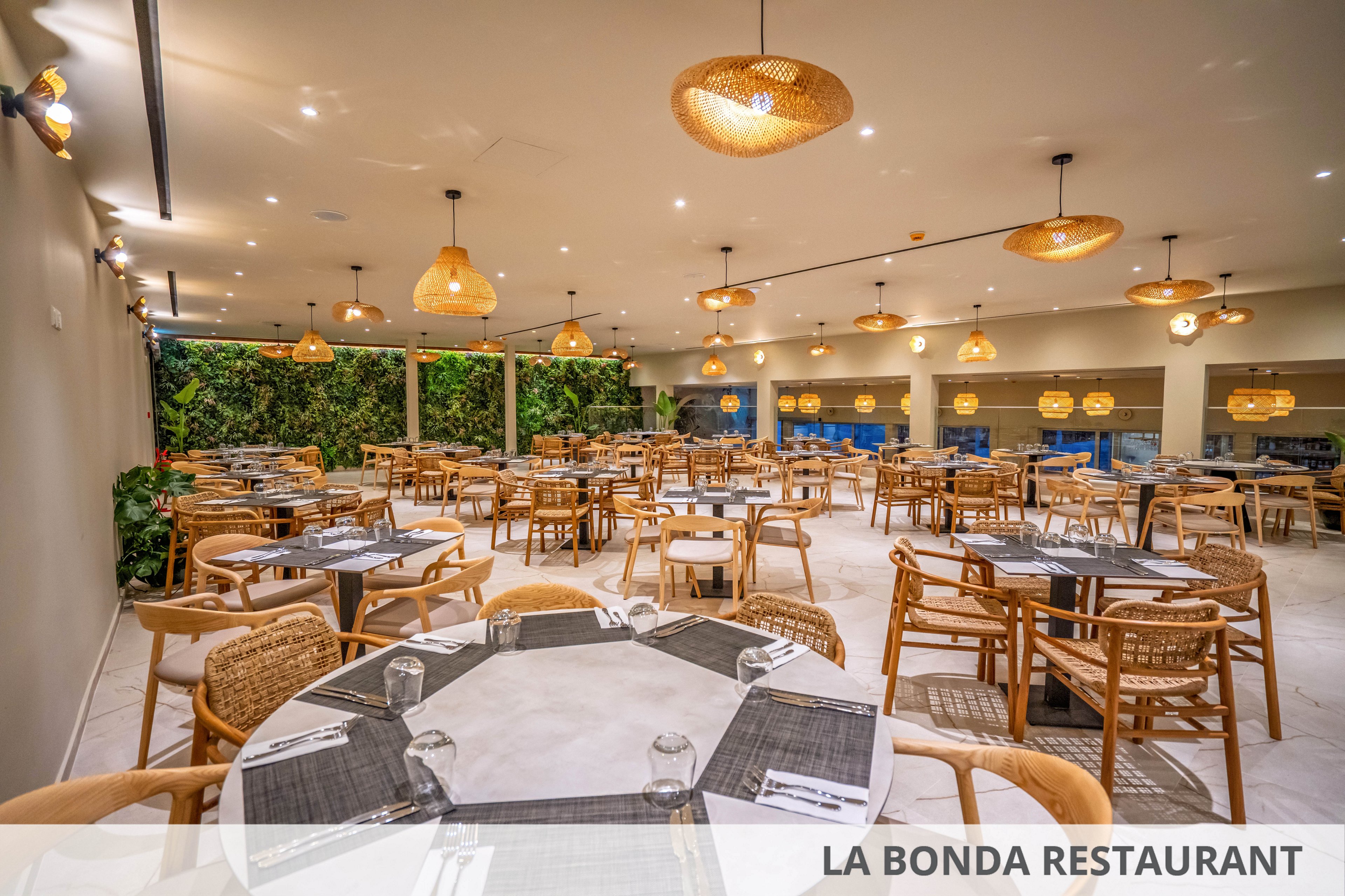 A spacious contemporary restaurant dining area is shown with neatly set round and rectangular tables. Wooden chairs and woven rattan accents create a warm, natural look throughout the room. Pendant lights hang from the ceiling, casting a soft glow over the neutral-toned floor and walls. Large windows and a green plant wall add depth and a relaxed, inviting atmosphere to the interior.