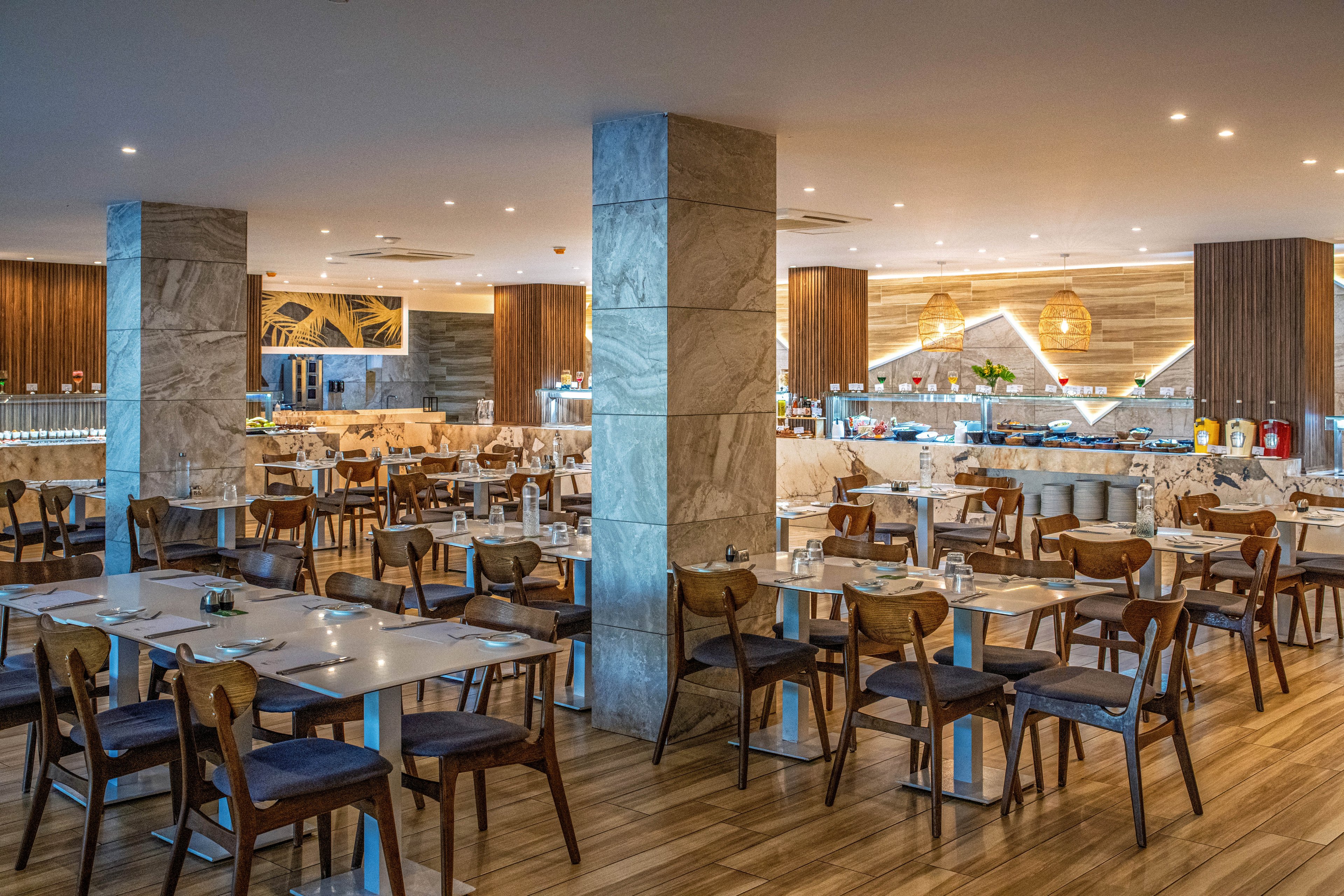 Spacious restaurant dining room with neatly arranged wooden tables and chairs. The setting features a contemporary buffet area in the background with warm pendant lighting and marble columns. Wooden flooring and neutral tones create a clean, modern atmosphere. No people are present, emphasizing the orderly layout and ambient lighting.