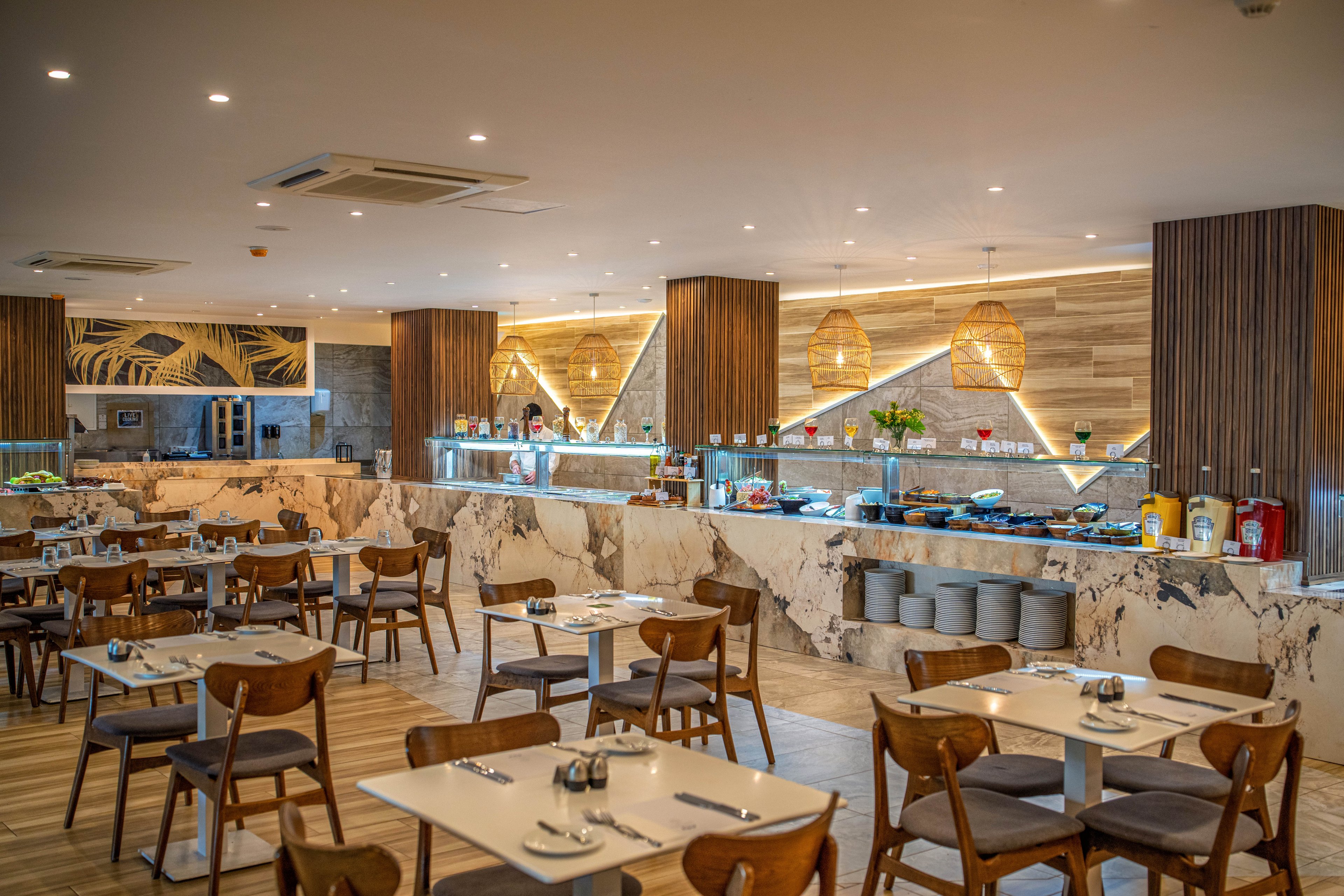 A contemporary buffet restaurant features neatly arranged wooden tables and chairs in the foreground. The back wall showcases a long marble counter with various food stations and beverage dispensers. Warm pendant lights, wood paneling, and decorative shelving create a cozy yet upscale atmosphere. The space appears clean, organized, and ready to welcome guests.
