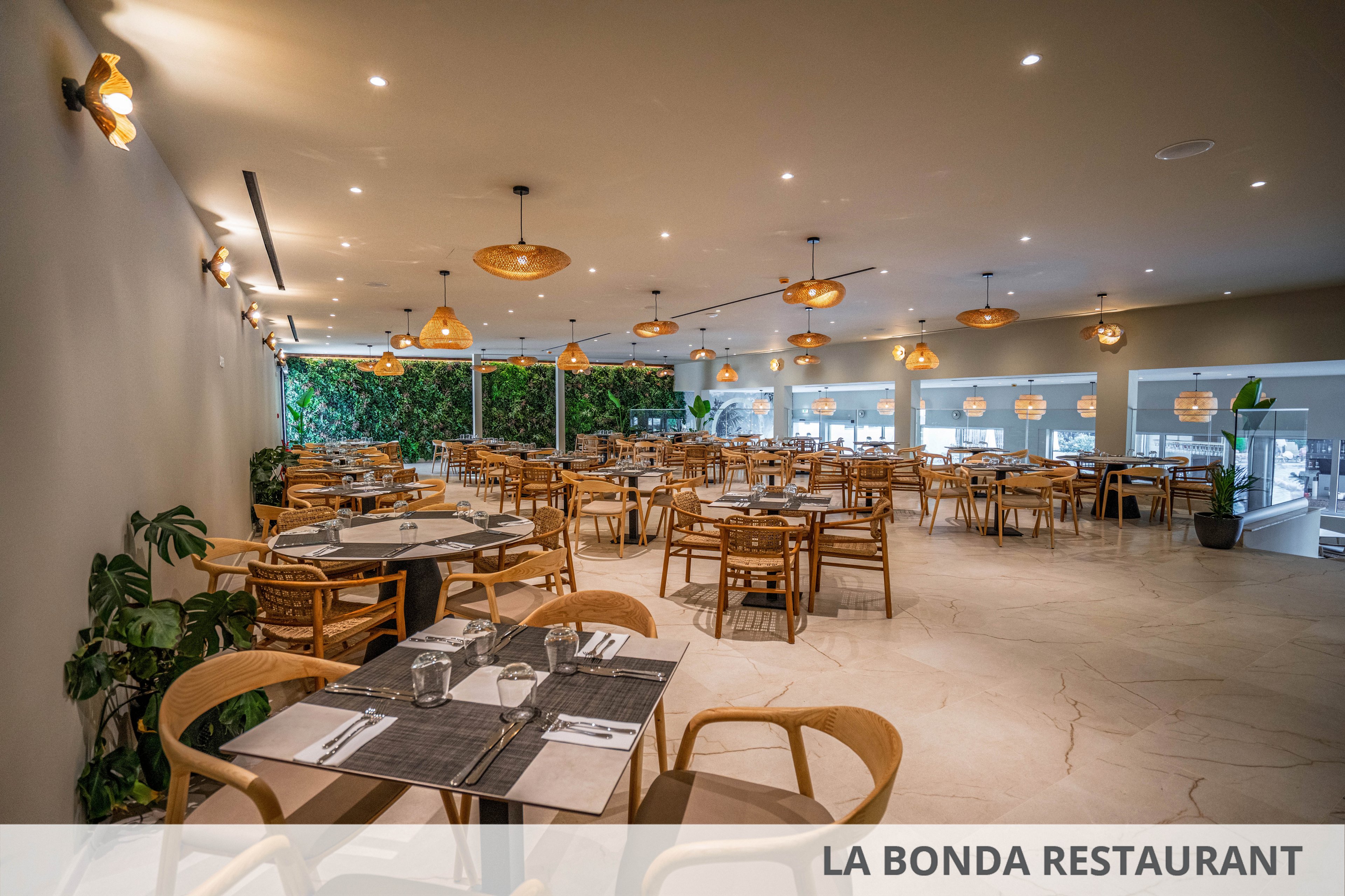 A spacious contemporary restaurant dining room is shown with neatly arranged wooden tables and chairs. Place settings with glasses, plates, and cutlery are prepared on dark placemats, ready for guests. Warm pendant lights hang from the ceiling, casting a cozy glow over the neutral-toned floor and walls. A green plant wall and potted plants add natural accents to the clean, minimalist interior.