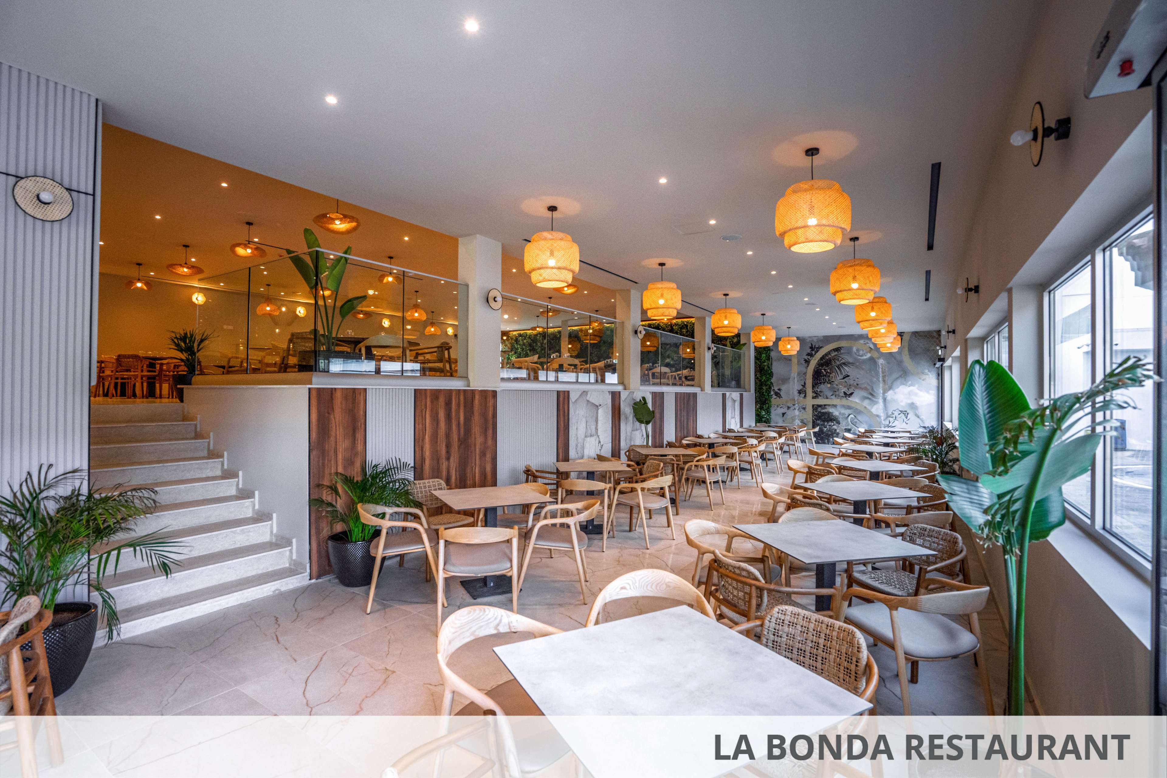 A spacious modern restaurant interior features neatly arranged wooden tables and chairs. Warm pendant lights hang from the ceiling, creating a cozy and inviting atmosphere. Large windows on the right bring in natural light, while green plants add freshness throughout the space. A raised mezzanine dining area is accessible by stairs, enhancing the open and contemporary design.