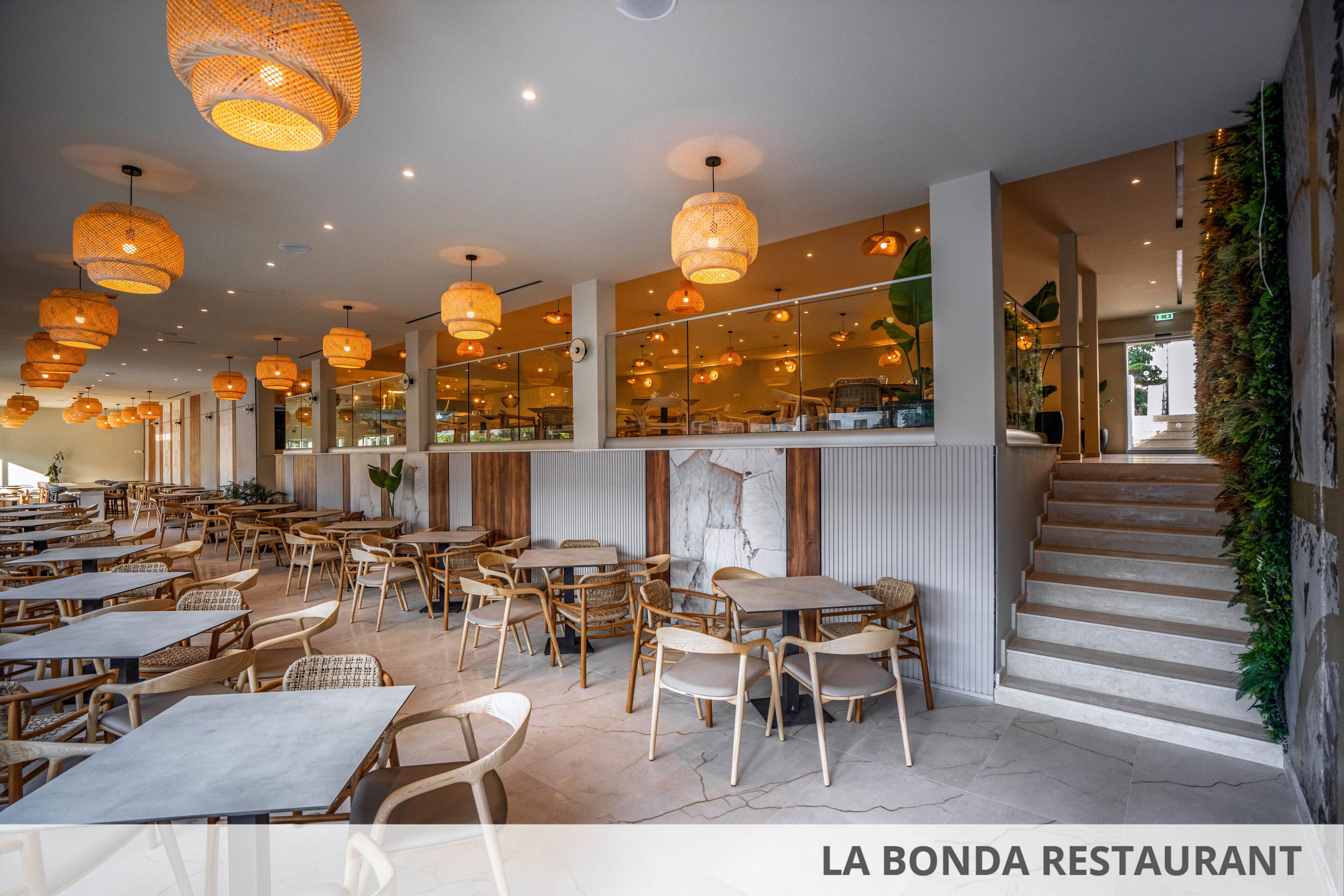A spacious contemporary restaurant interior features rows of wooden tables and chairs arranged neatly in an open dining area. Warm woven pendant lights hang from the ceiling, casting a soft glow over the neutral-toned space. Large windows and glass partitions separate the dining room from the service area, while a staircase leads up to another level. Green plants and natural materials add a relaxed, inviting atmosphere to the modern design.