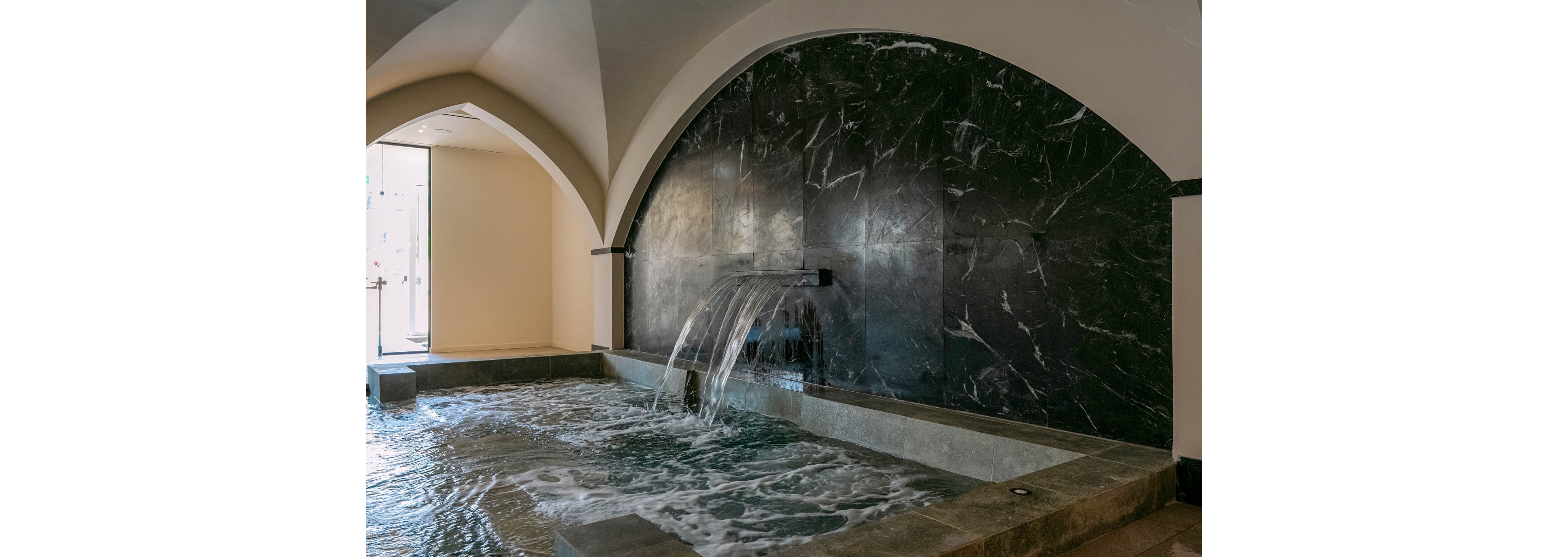 An indoor spa pool features a wide waterfall spout pouring water into a rectangular basin. The pool is framed by stone edges and set against a dark marble wall with a large arched recess. Gentle ripples and bubbles on the water surface suggest movement and relaxation. Soft natural light enters from the side, enhancing the tranquil, luxurious atmosphere of the spa interior.
