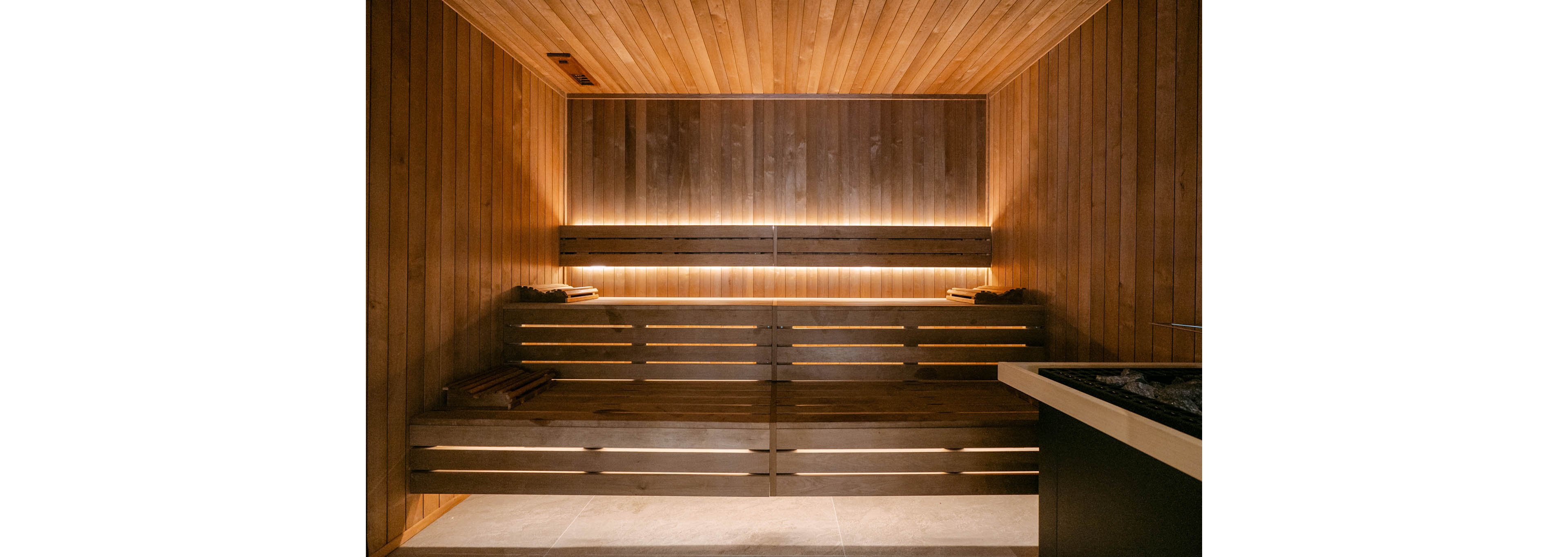 A contemporary sauna interior features smooth wooden walls, ceiling, and tiered bench seating. Warm, indirect lighting runs along the back wall, creating a cozy and inviting atmosphere. A stone-filled heater unit is visible in the foreground, emphasizing the spa and wellness setting. The space appears clean, minimal, and designed for relaxation and comfort.