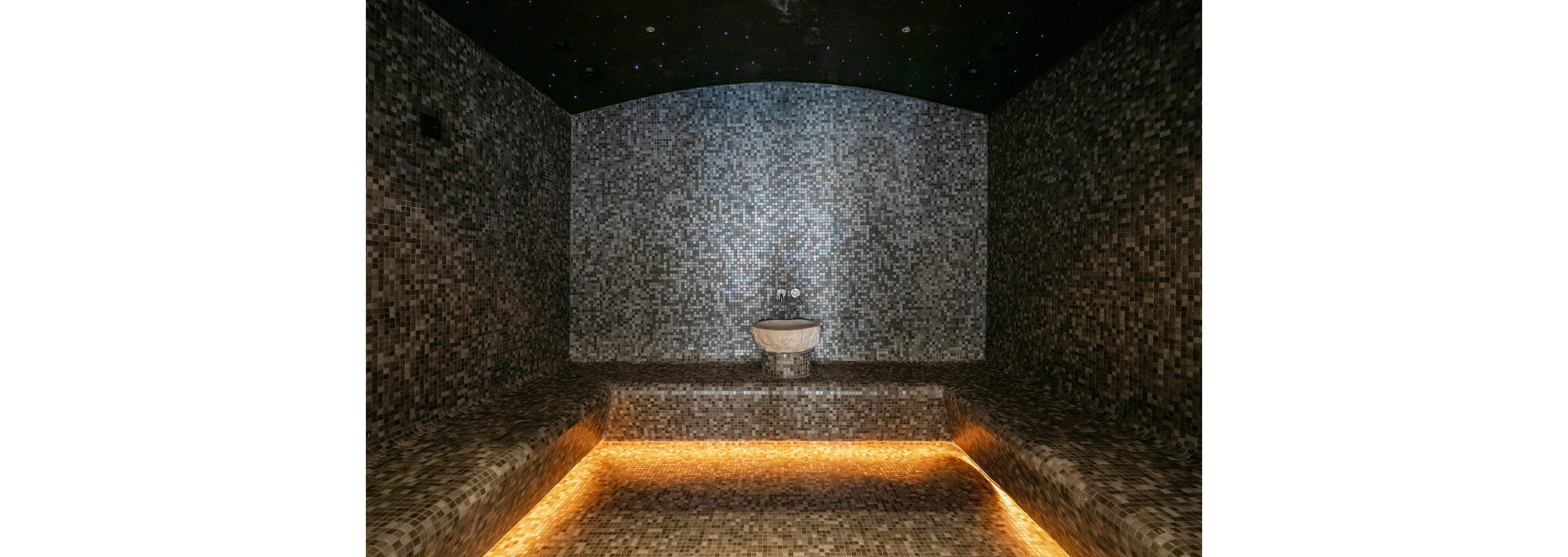 A contemporary steam room is fully covered in small mosaic tiles, creating a textured and immersive environment. A white basin is centered against the back wall, serving as a focal point in the space. Warm yellow lighting runs beneath the seating ledge, casting a soft glow across the tiled surfaces. The dark ceiling above is dotted with tiny lights, evoking a starry night effect and enhancing the relaxing atmosphere.
