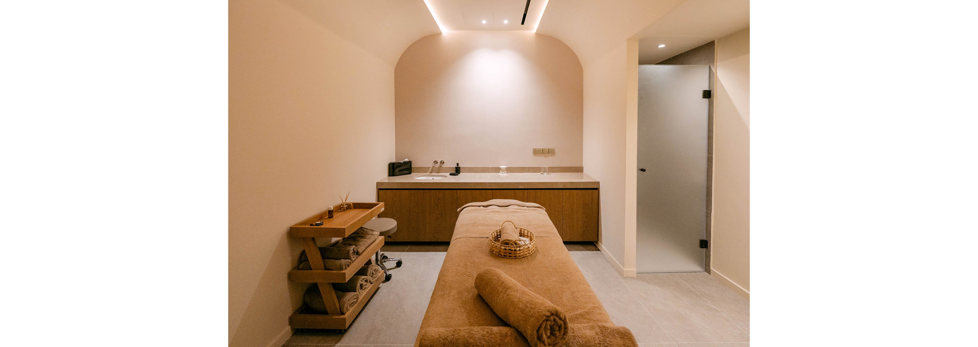 A warm, softly lit spa treatment room features a central massage table covered in beige towels. A wicker basket and neatly rolled towel sit at the end of the table, ready for a relaxing session. Wooden shelves on the left hold spa accessories and products, while a long counter with sink and storage runs along the back wall. Minimalist design, neutral colors, and gentle lighting create a calm, inviting atmosphere.