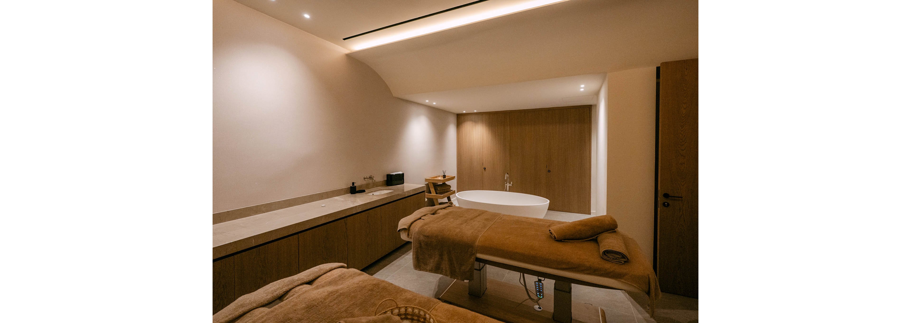 A warm, minimalist spa treatment room features two massage tables covered with soft brown towels. A freestanding white bathtub stands against a wooden wall, adding a touch of luxury to the space. Built-in cabinetry runs along one side with neatly arranged accessories, under soft recessed ceiling lights. The overall ambiance is calm and inviting, emphasizing relaxation and wellness.