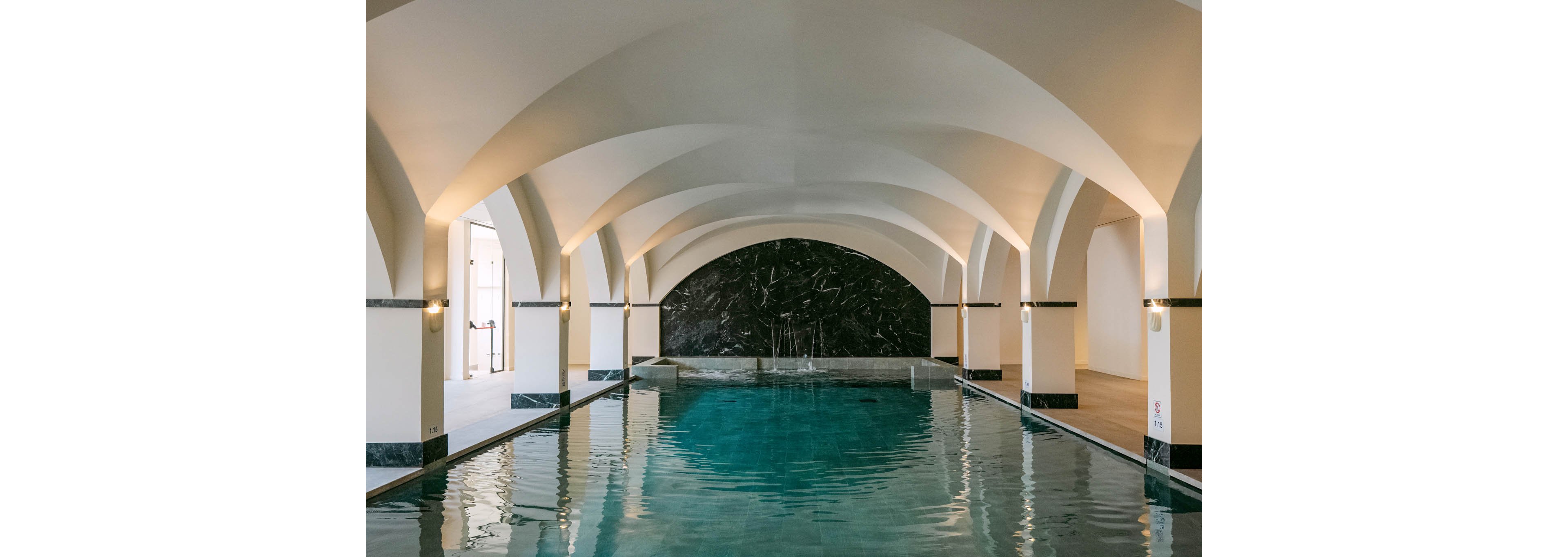 An elegant indoor swimming pool is set beneath a series of white arched ceilings. Soft wall lights reflect on the calm turquoise water, creating a serene atmosphere. A dark marble wall forms a dramatic focal point at the far end of the pool. The space appears quiet and refined, suggesting a spa or upscale wellness retreat.