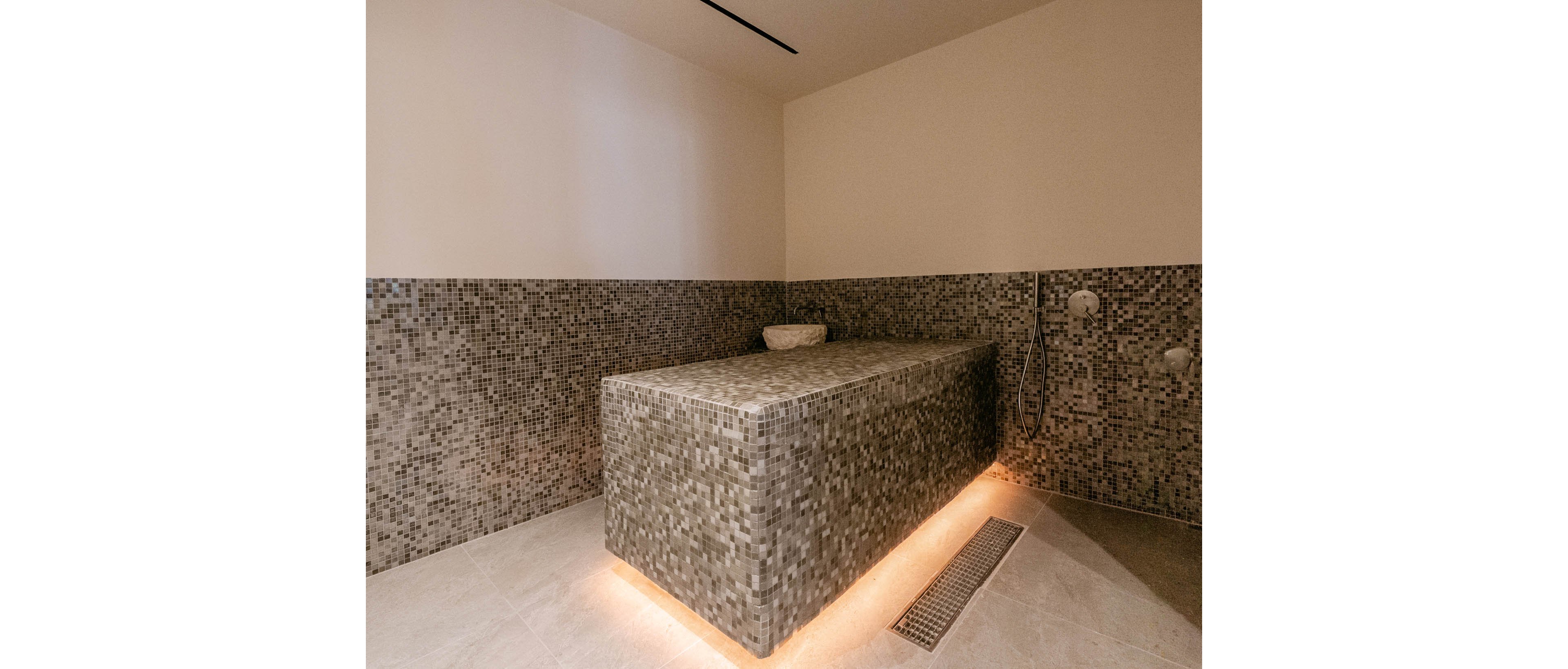 A contemporary spa treatment room features a large rectangular tiled massage or steam table as the central element. The walls and table are covered in small mosaic tiles, creating a cohesive, textured look. Warm underlighting around the base of the table adds a relaxing ambiance to the neutral-toned space. A wall-mounted shower fixture and floor drain suggest a wet-room or hammam-style environment.