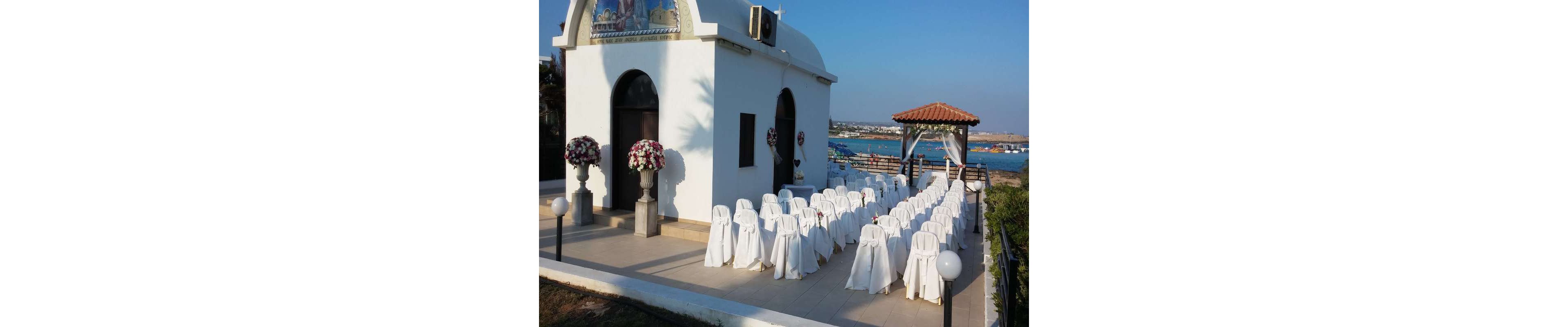 An outdoor wedding ceremony setup is arranged beside a small white chapel overlooking the sea. Rows of white chairs with covers and bows face a floral-decorated entrance and a pergola-style gazebo. Large flower arrangements in shades of white and deep red flank the chapel doorway. The bright blue sky and distant coastline create a serene coastal backdrop. The scene suggests a formal, romantic event in a Mediterranean-style setting.