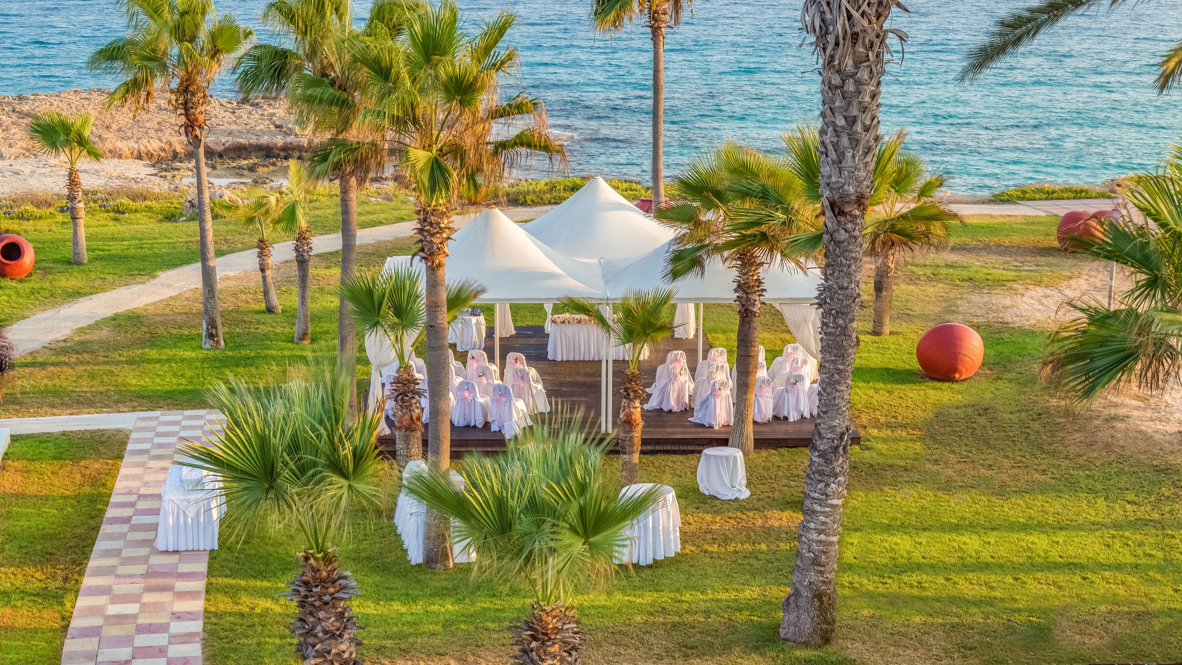 An elegant outdoor event setup is arranged under a white canopy surrounded by palm trees near the sea. White-covered chairs and tables are placed neatly on a wooden deck, suggesting a wedding or formal celebration. The lush green lawn, stone pathway, and large red decorative spheres add visual interest to the coastal setting. Calm blue water and rocky shoreline in the background create a serene, tropical atmosphere.