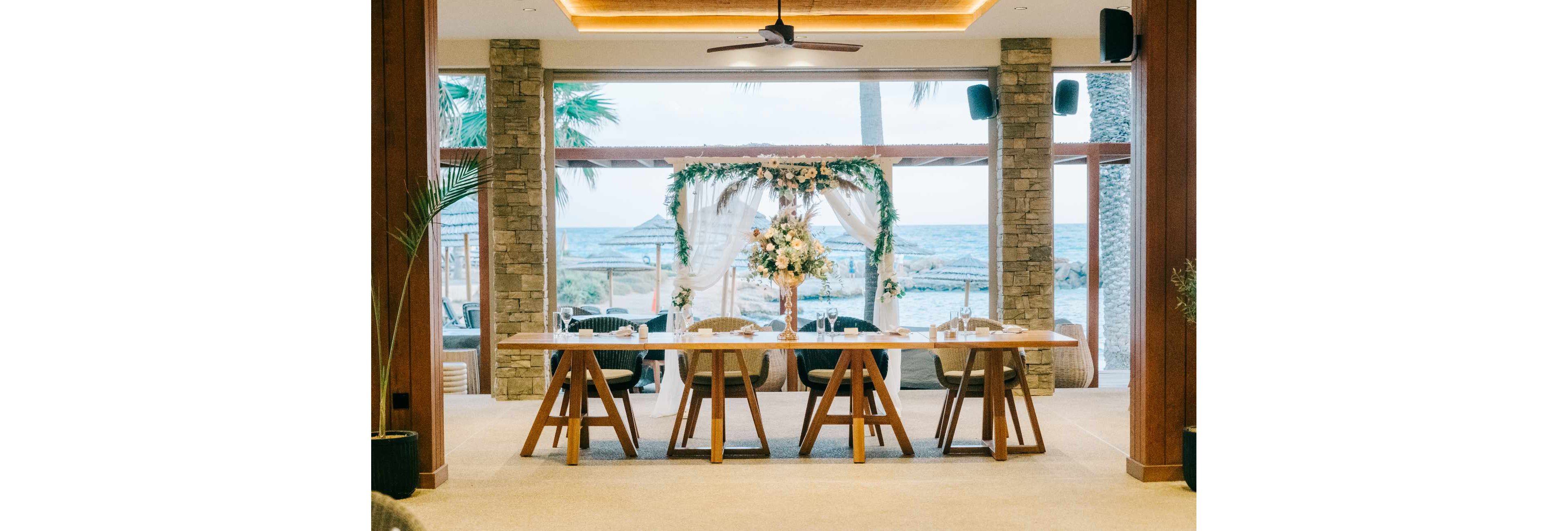 A long wooden table is beautifully arranged for a wedding reception in an open, airy venue. Floral arrangements and greenery decorate an archway framing a view of the ocean and palm trees outside large glass windows. Neutral tones, soft lighting, and natural materials create a relaxed yet sophisticated coastal atmosphere. The scene suggests a formal event or celebration by the sea.