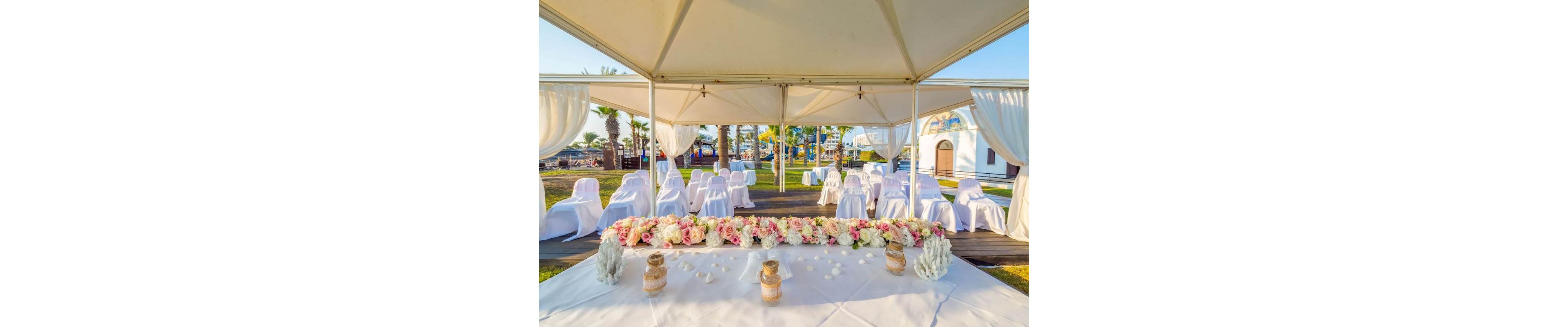 An elegant outdoor wedding ceremony is arranged under a white canopy near the seaside. Rows of white-covered chairs face a long table adorned with pink and white floral arrangements. Decorative candles and small ornaments sit on the table, enhancing the romantic atmosphere. Palm trees, green lawn, and distant water views create a bright, sunny backdrop for the event.