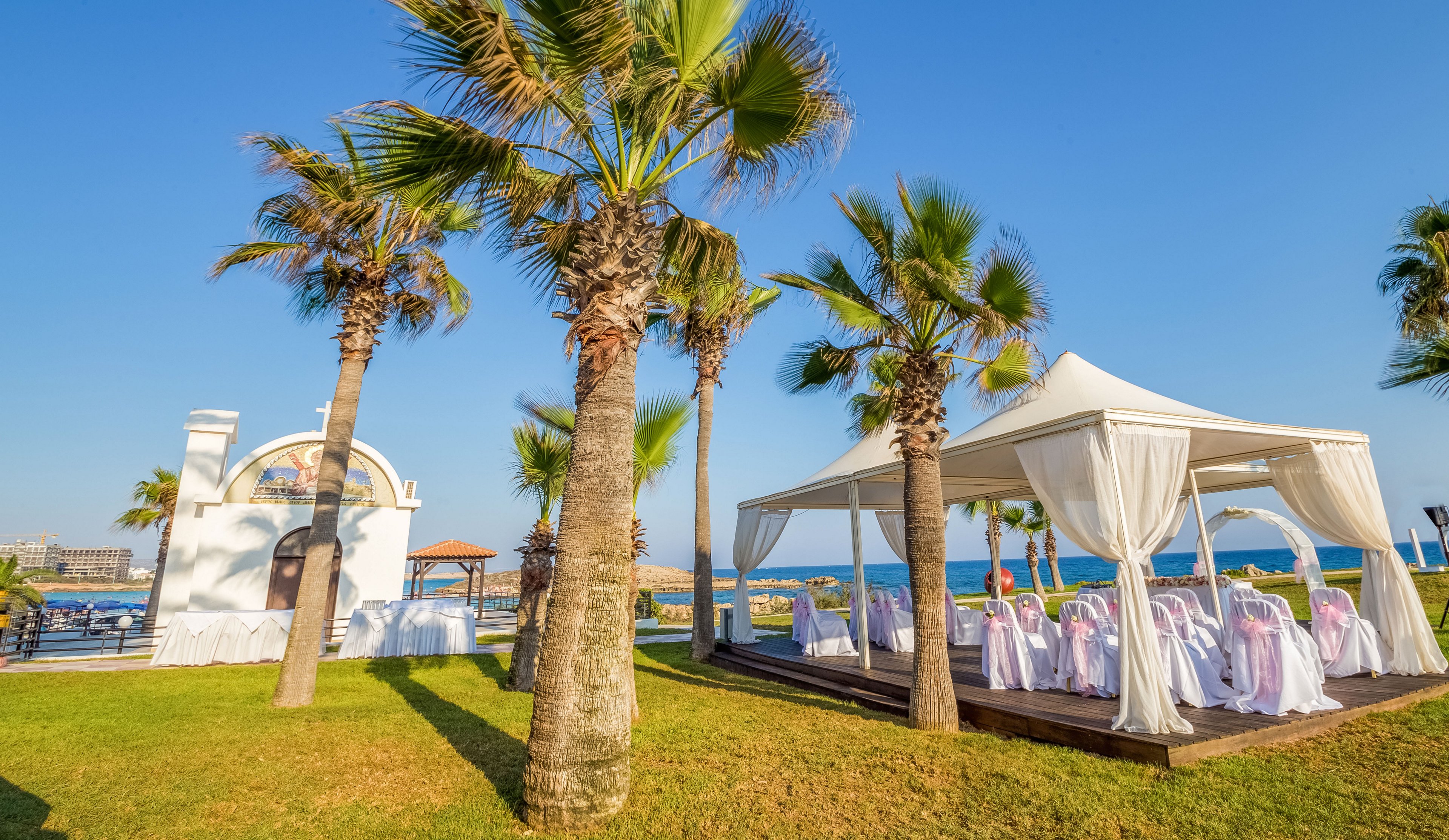 An elegant outdoor wedding setup is arranged on a grassy lawn beside the sea. A white canopy tent shelters rows of chairs draped in white covers and pink sashes. Palm trees frame the scene, and a small white chapel-like structure stands nearby. The bright blue sky and ocean create a sunny, romantic coastal atmosphere.
