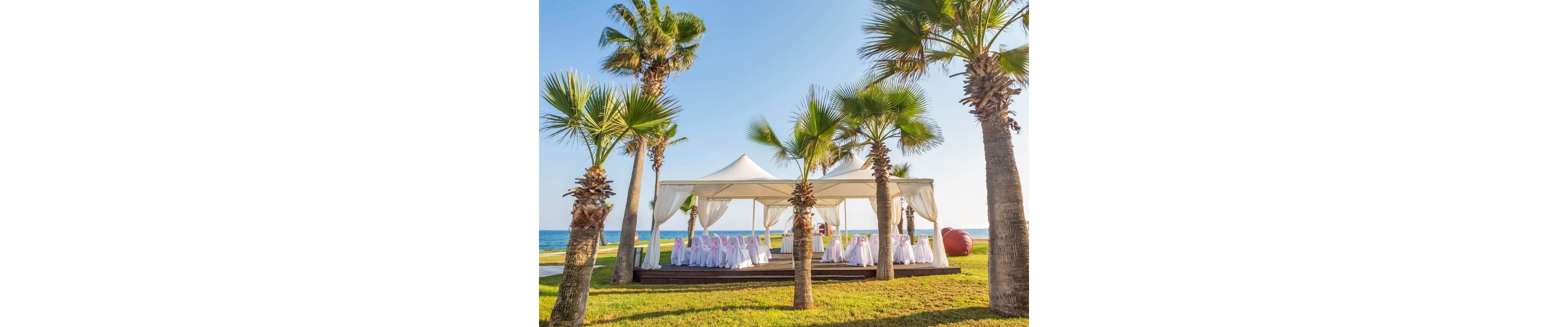 An elegant white canopy is set up on a wooden deck near the sea, prepared for a formal event or wedding reception. Rows of chairs draped in white covers with pink sashes are arranged neatly beneath the tent. Tall palm trees surround the structure, framing the view of the blue ocean and clear sky. The bright daylight and lush green grass create a warm, festive atmosphere in a coastal setting.