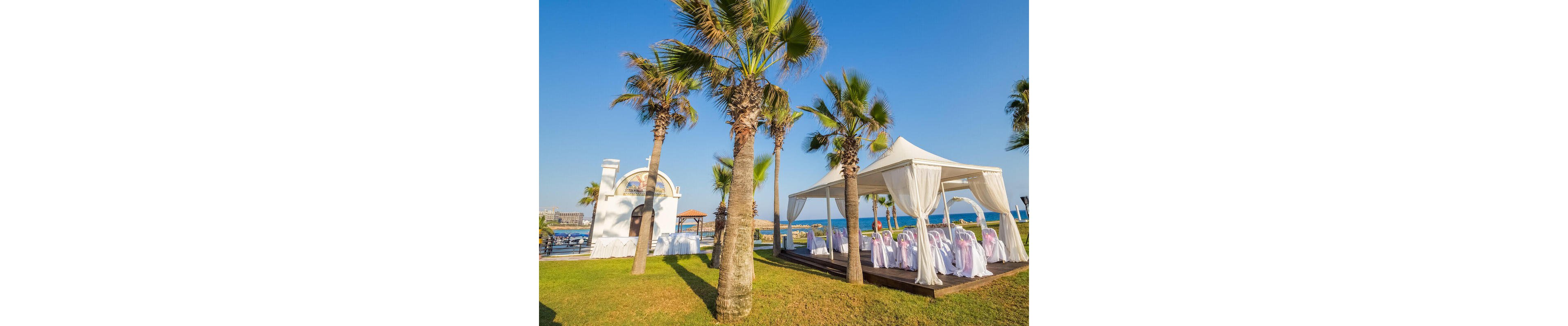An elegant outdoor wedding venue is arranged on a grassy lawn beside the sea. A white canopy tent with flowing curtains shelters rows of chairs draped in white covers and pastel bows. Palm trees surround the setup, adding a tropical feel under a clear blue sky. In the background, a small white structure and distant buildings frame the coastal setting, creating a bright and festive atmosphere.