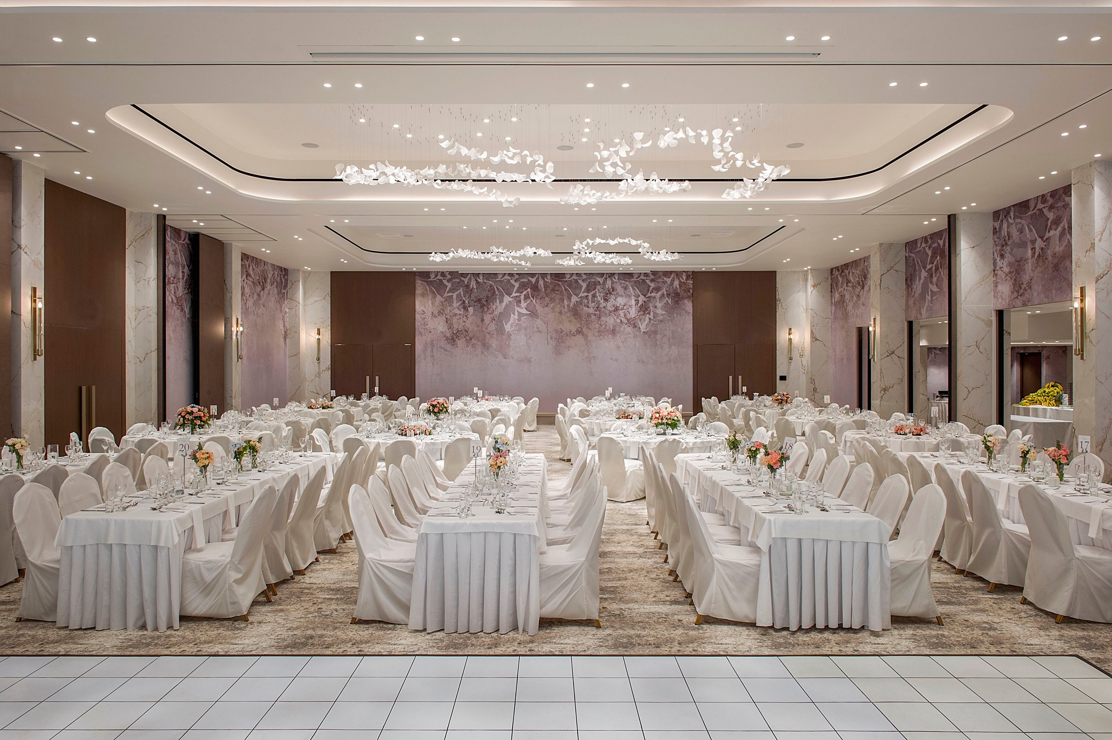 A spacious banquet hall is arranged with long rows of tables covered in white linens and matching white chair covers. Floral centerpieces and neatly arranged table settings suggest a formal celebration or reception. Soft, modern ceiling lighting and decorative wall panels create a sophisticated atmosphere. The room appears ready to host a large gathering in a refined, contemporary setting.