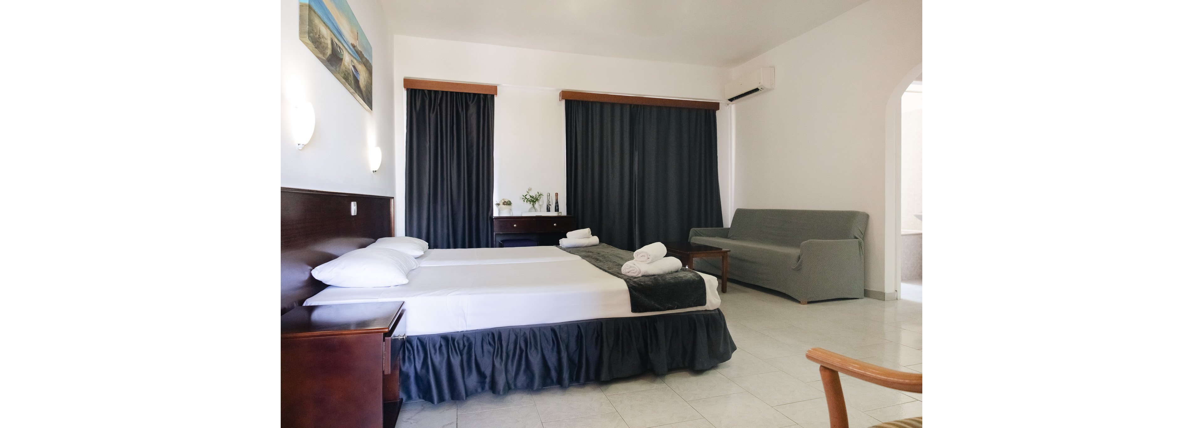 A clean, spacious hotel room features a large double bed with white linens and neatly folded towels. Dark curtains cover the windows, contrasting with the white walls and tiled floor. A gray sofa sits against the wall near a small wooden table, and an air conditioning unit is mounted above. Minimal decor and soft lighting create a simple, comfortable atmosphere suitable for guests.