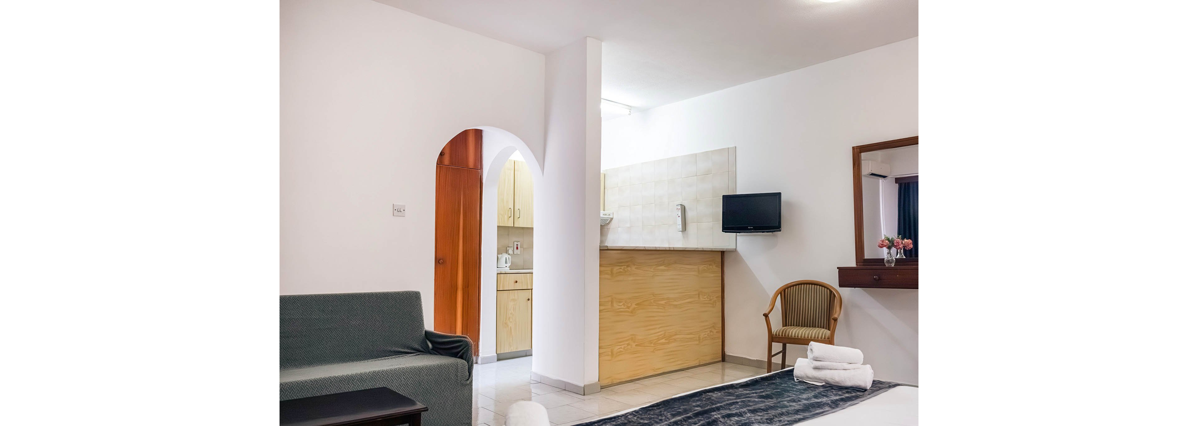 Bright contemporary hotel room featuring a bed in the foreground and a small seating area. A compact kitchenette is visible through an arched doorway with light wood cabinets and countertop. A wall-mounted flat screen television faces the bed above a wooden panel. The space is decorated in neutral white tones with simple furniture and a vase of flowers adding color.