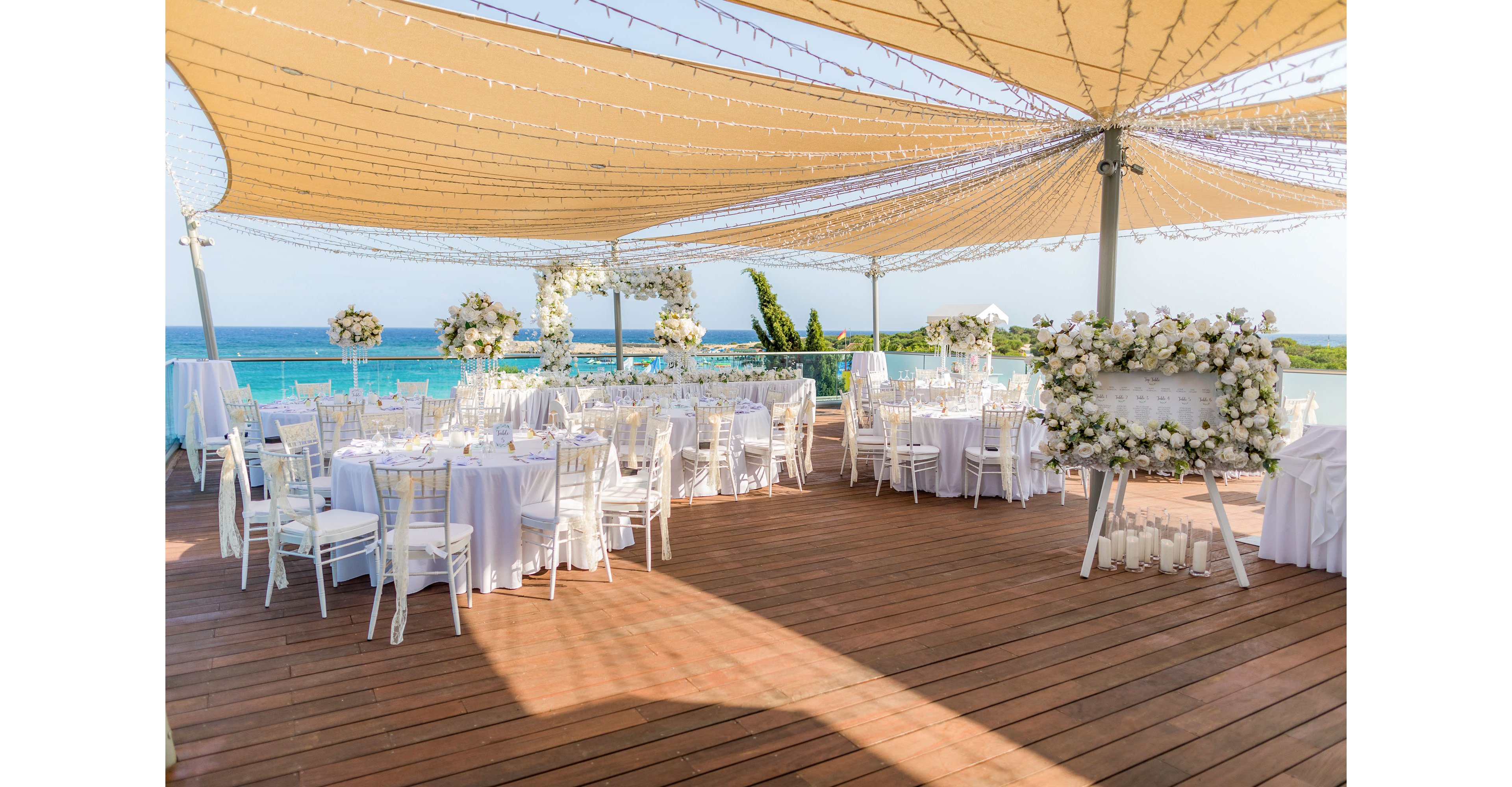 An elegant wedding reception is arranged on a wooden terrace overlooking a bright blue sea. Round tables with white linens and white chairs are neatly organized under large beige shade canopies. Floral arrangements of white flowers decorate the tables, arch structures, and a large display at the front. String lights hang across the canopy, adding a festive touch to the open-air venue. The scene is calm, romantic, and ready for guests to arrive.