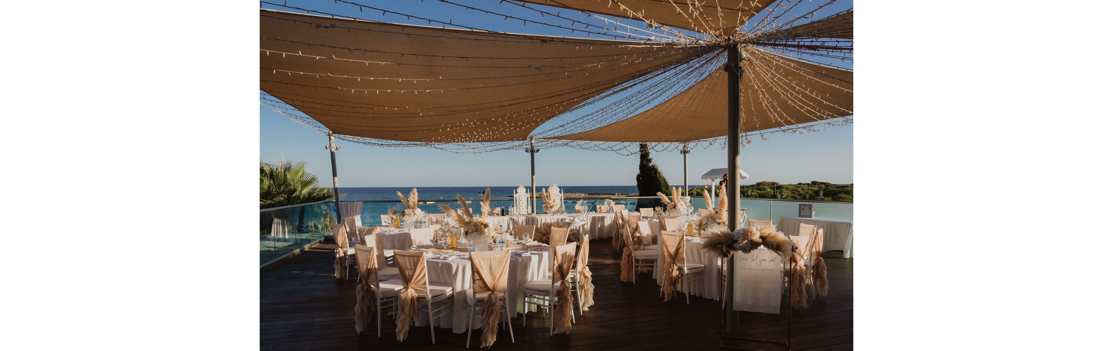 An outdoor wedding reception is arranged on a wooden terrace overlooking the sea. Round tables with white linens and chairs draped in beige fabric are decorated with floral centerpieces and candles. A large canopy with string lights provides shade and creates a festive atmosphere. The coastal backdrop, clear sky, and soft neutral decor give the scene a romantic, sophisticated feel.