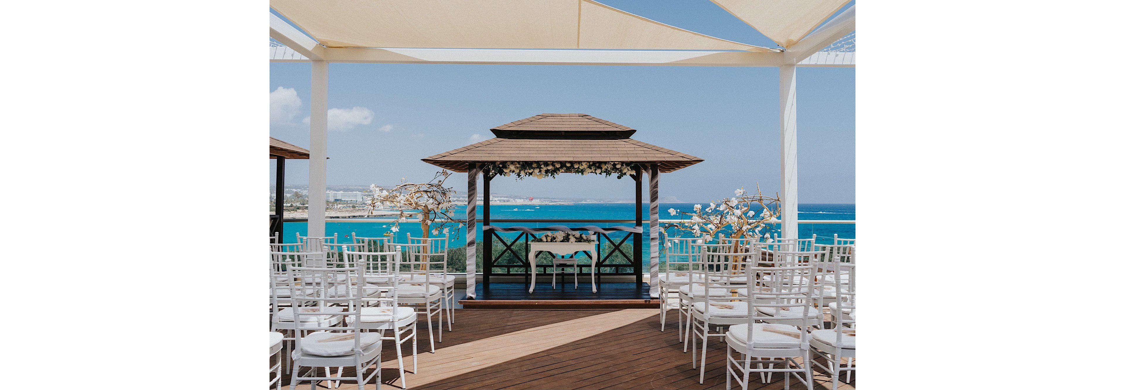 An elegant wooden gazebo is prepared for a wedding ceremony overlooking a bright blue sea. White chairs are arranged in neat rows on a wooden deck, facing the decorated altar table under the canopy. Floral arrangements and draped greenery adorn the gazebo, adding a romantic touch to the coastal setting. Sun shades above create soft, diffused light, enhancing the serene and festive atmosphere.