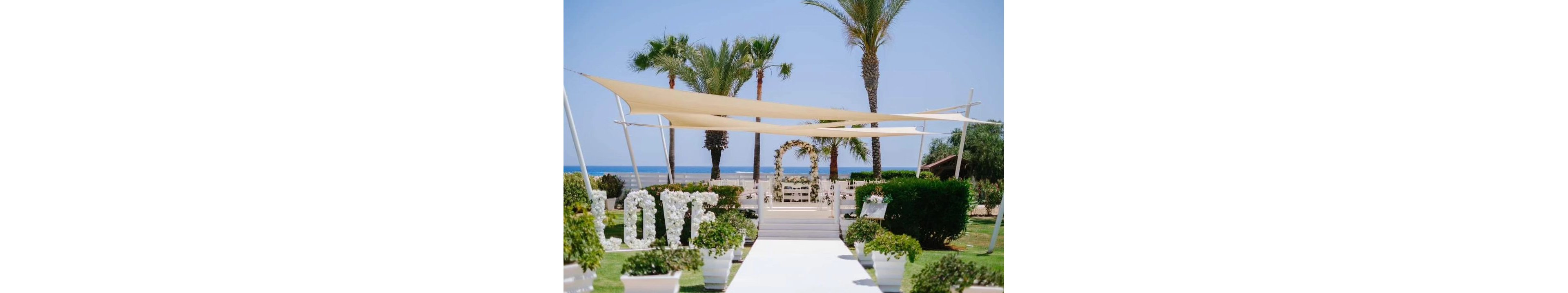An elegant outdoor wedding aisle is set up in a garden overlooking the sea. White floral arrangements and potted greenery line the white walkway leading to an arch decorated with flowers. Light fabric canopies provide shade above the aisle, while palm trees frame the view of the blue ocean and sky. Large white letters spelling the word LOVE stand prominently near the path, enhancing the romantic atmosphere.