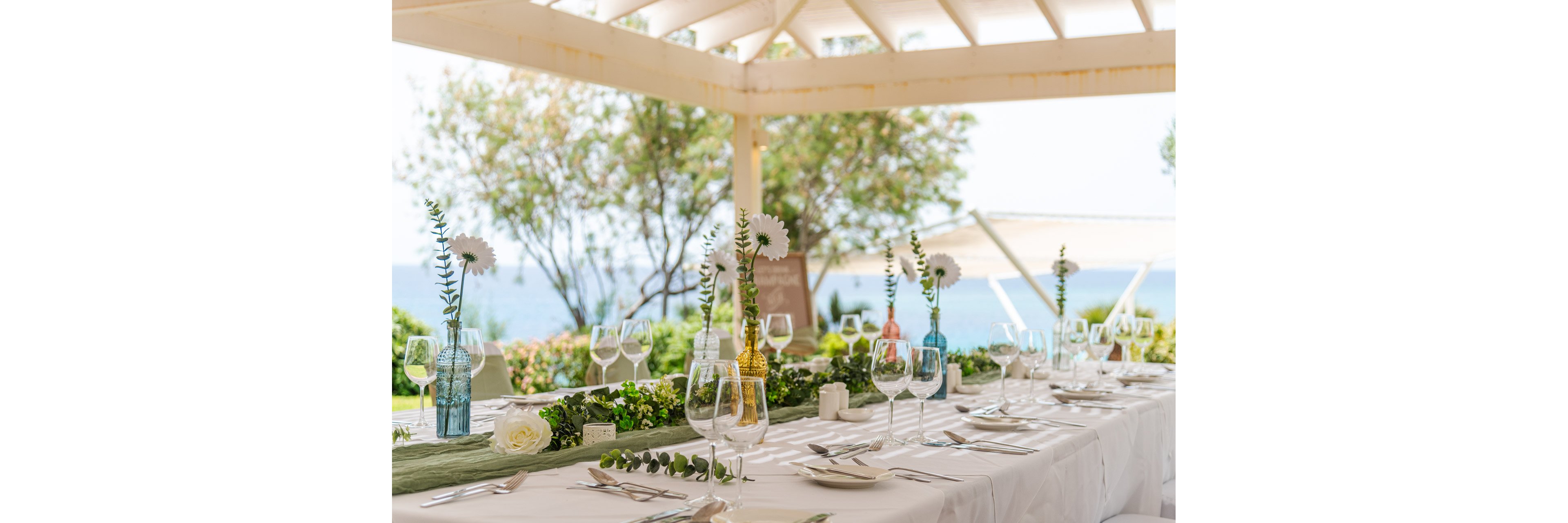 A long banquet table is set for an elegant event under a bright open pavilion by the sea. White tablecloths, glassware, and neatly arranged plates create a refined dining setting. Colorful glass vases with white flowers and greenery run along the center of the table as a decorative centerpiece. Soft natural light and a view of the ocean in the background suggest a relaxed, romantic celebration.