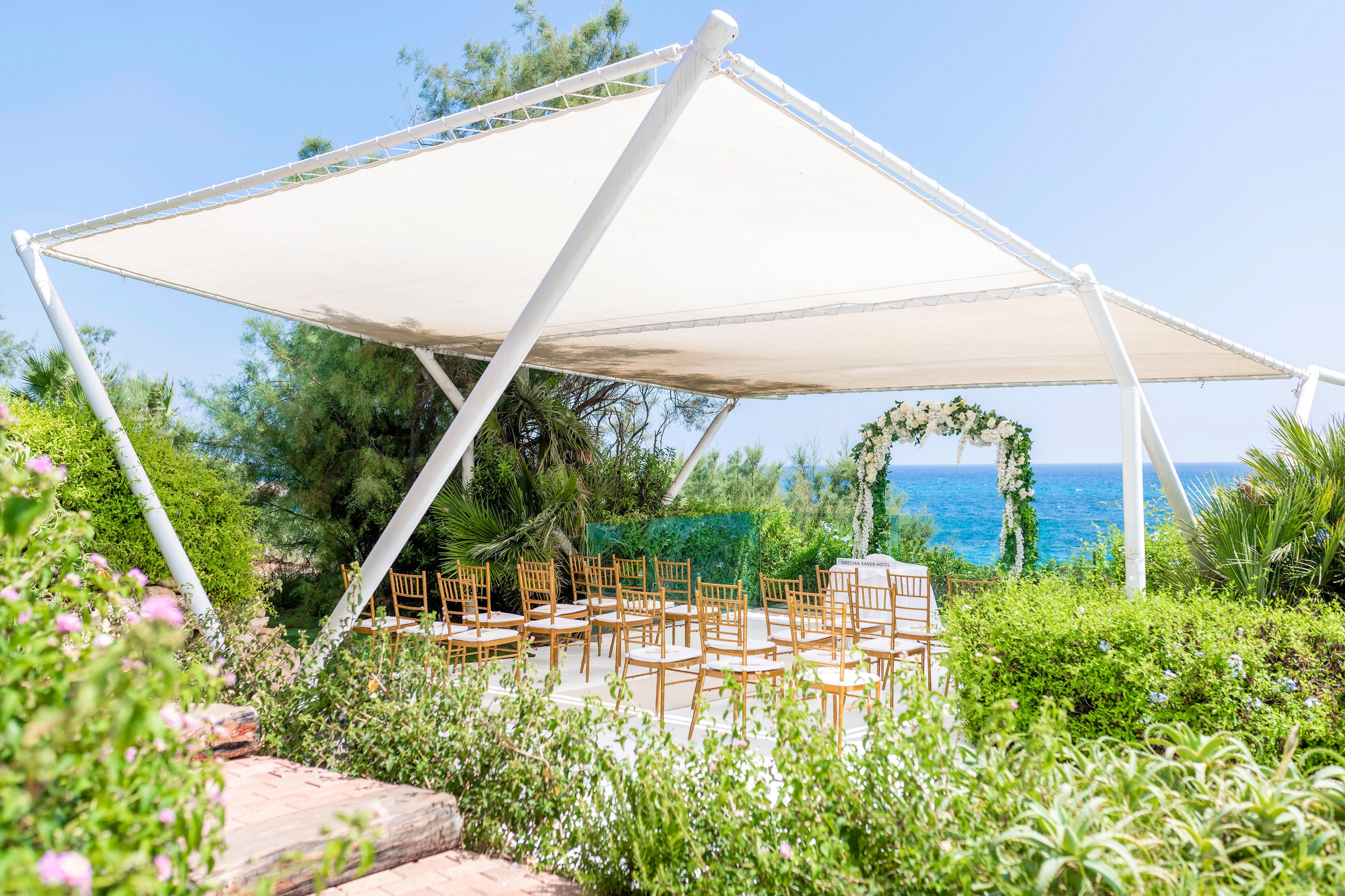 An outdoor wedding ceremony setup is arranged beneath a modern white canopy. Wooden chairs are neatly aligned facing a floral arch that frames a view of the blue sea. Lush green plants and shrubs surround the seating area, creating a natural garden atmosphere. The bright daylight and clear sky give the scene a fresh, romantic, and summery feel.
