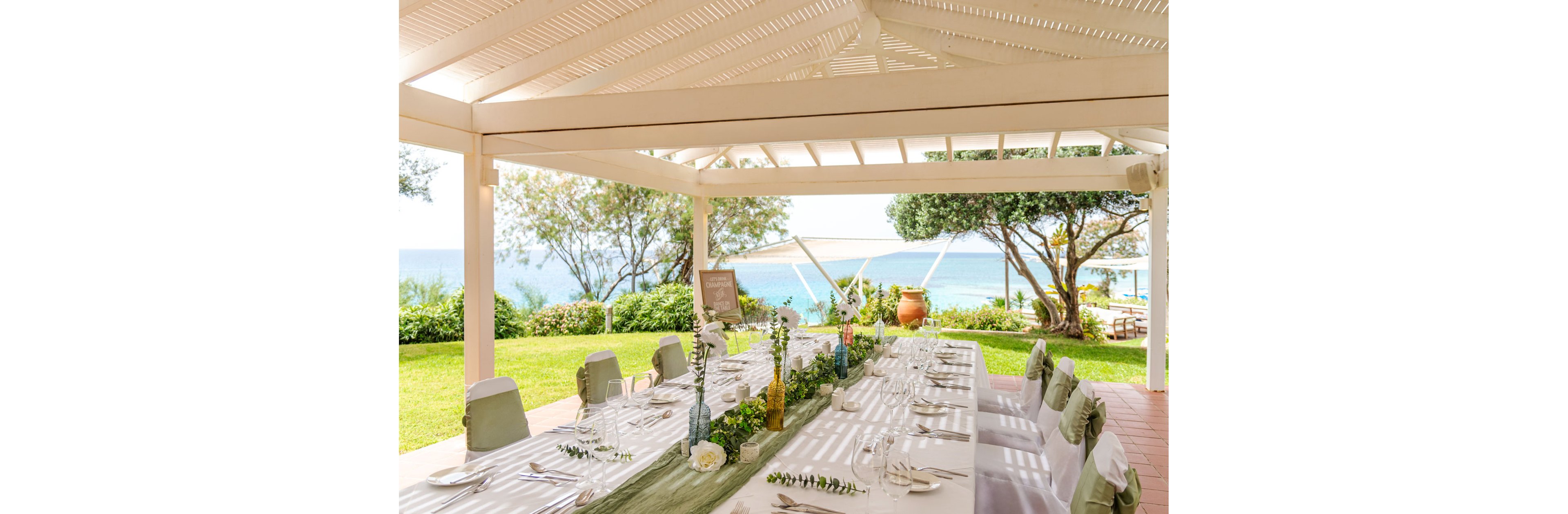 A long, elegantly set dining table is arranged under a white pergola overlooking the sea. The table is decorated with white linens, green runners, and floral centerpieces, creating a refined celebration setting. Soft natural light and greenery surround the space, with trees, lawn, and terracotta pots enhancing the coastal atmosphere. The scene suggests a formal event such as a wedding reception or special celebration in a serene seaside garden.