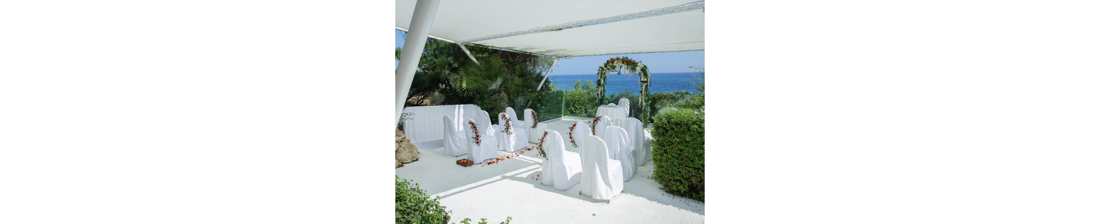 An intimate wedding ceremony setup is arranged under a white canopy overlooking the sea. White chairs draped in covers form two rows facing a floral arch decorated with greenery and orange accents. The bright setting is surrounded by lush bushes and palm trees, with the blue ocean visible in the background. The scene suggests a calm, romantic atmosphere for a coastal celebration.