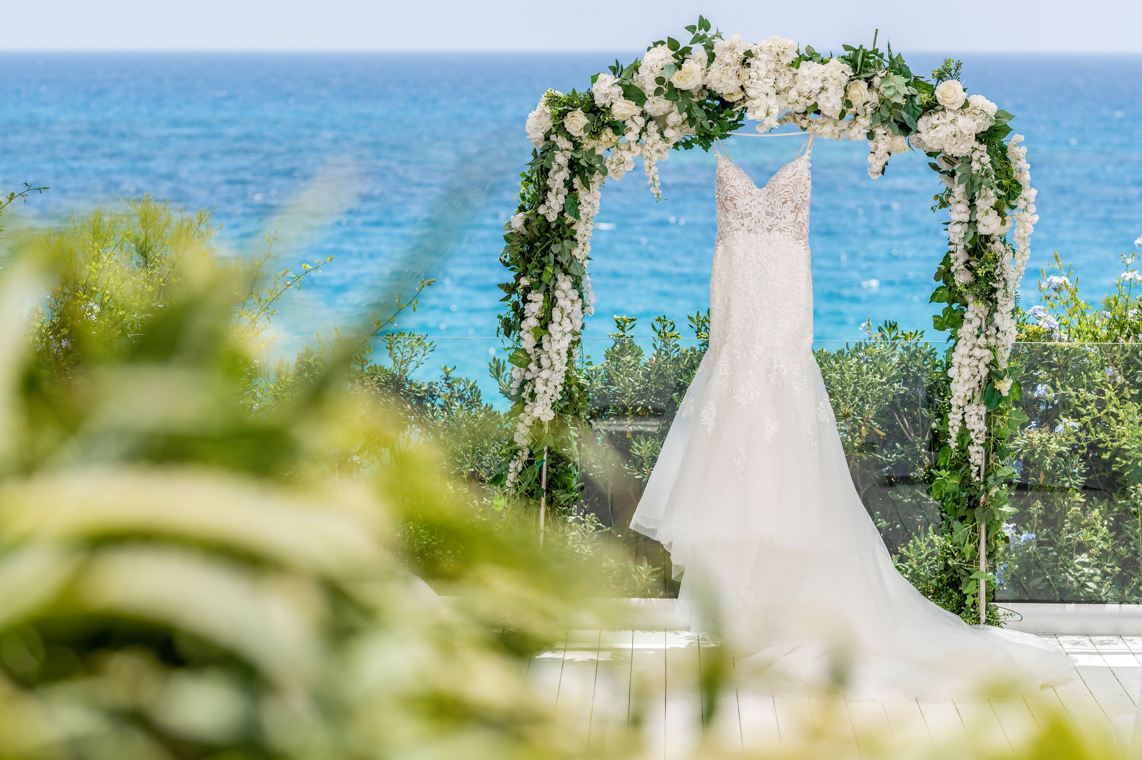A white lace wedding dress hangs gracefully from a floral arch overlooking a bright blue sea. The arch is decorated with lush green foliage and white flowers, framing the gown against the ocean backdrop. Sunlight highlights the delicate details of the dress and the soft pastel tones of the scene. Blurred greenery in the foreground adds depth and a romantic, outdoor atmosphere.