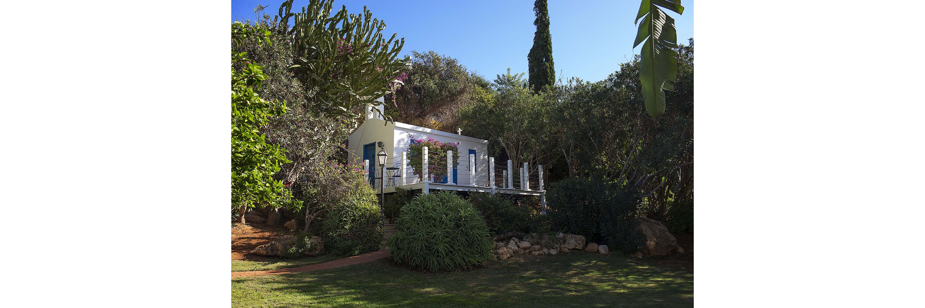 A small white house with blue doors and windows sits elevated on a wooden deck in a lush garden. The structure is partially hidden by dense trees, shrubs, and flowering plants. A narrow path and a classic lamp post lead toward the house, adding charm to the scene. Bright sunlight and deep shadows create a tranquil, secluded atmosphere.