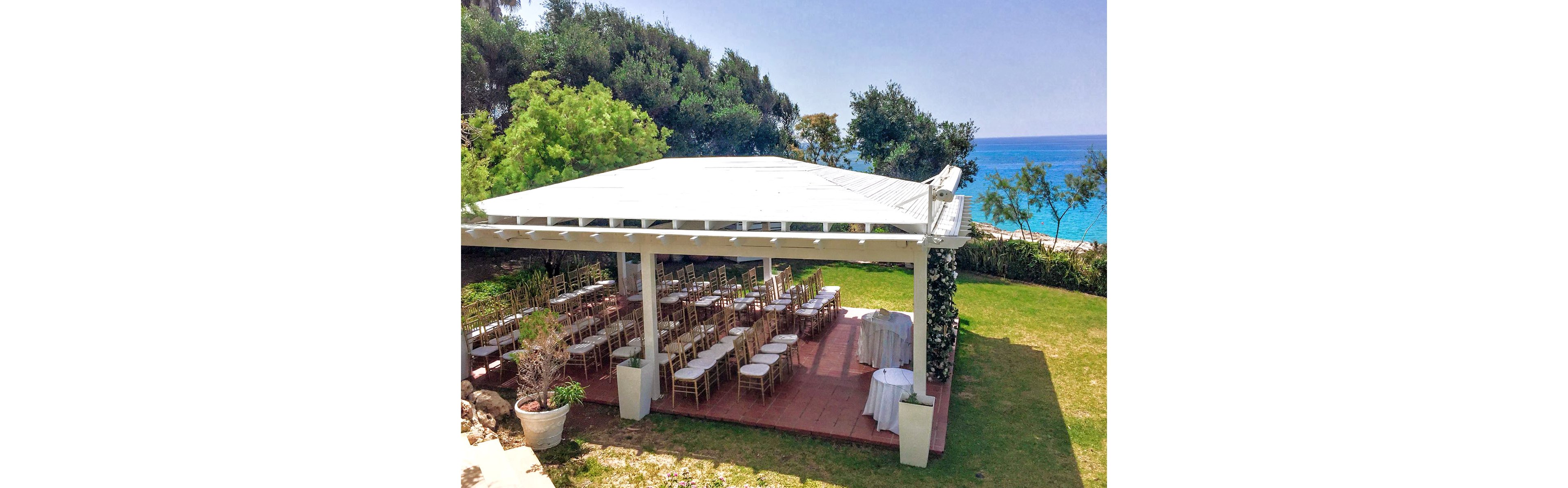 An outdoor wedding ceremony area is arranged under a white pergola near the sea. Rows of wooden chairs are neatly aligned facing the front, suggesting a formal event or celebration. The pavilion stands on a red-tiled platform surrounded by green grass and lush trees. In the background, bright blue ocean water and clear sky create a vibrant coastal atmosphere.