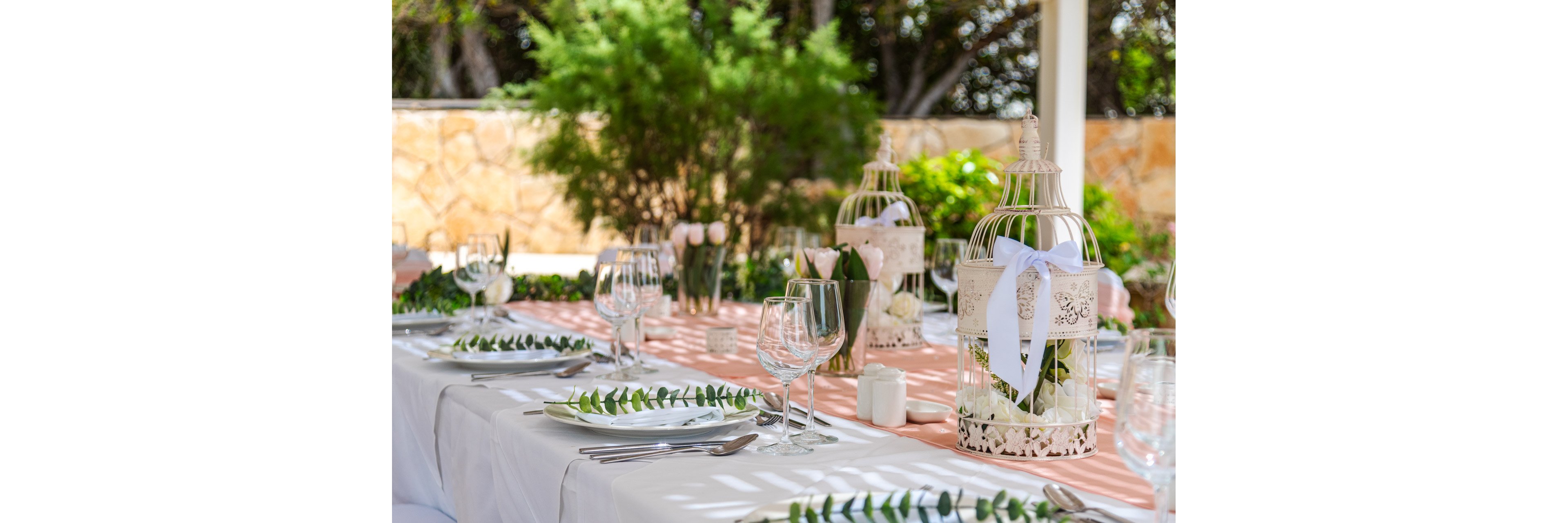 A long white-clothed table is set for an outdoor reception with elegant glassware and cutlery. Decorative white birdcages with ribbons and flowers serve as centerpieces along the table. Greenery garlands and simple leaf sprigs accent each place setting, adding a fresh natural touch. The scene is bright and airy, with soft pastel tones and a garden backdrop suggesting a wedding or special celebration.