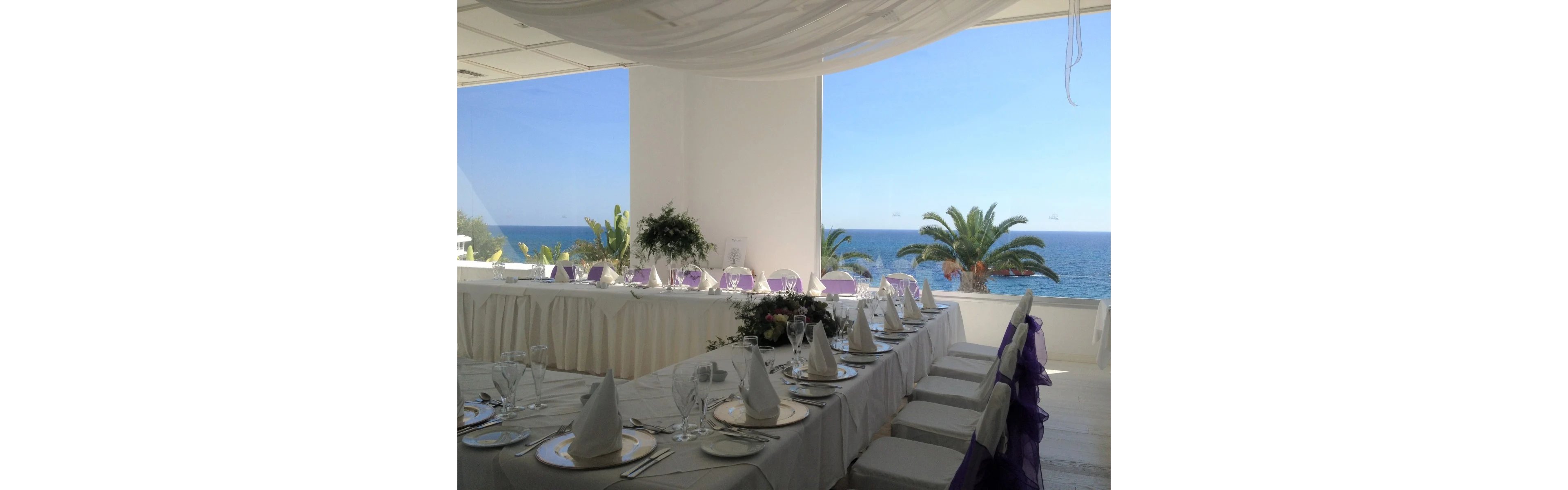 A long banquet table is elegantly set for a wedding reception in a bright seaside venue. White linens, folded napkins, and polished glassware create a refined and formal atmosphere. Purple chair sashes and floral centerpieces add soft color accents to the predominantly white decor. Large windows reveal a clear view of the blue ocean, palm trees, and a sunny sky outside.