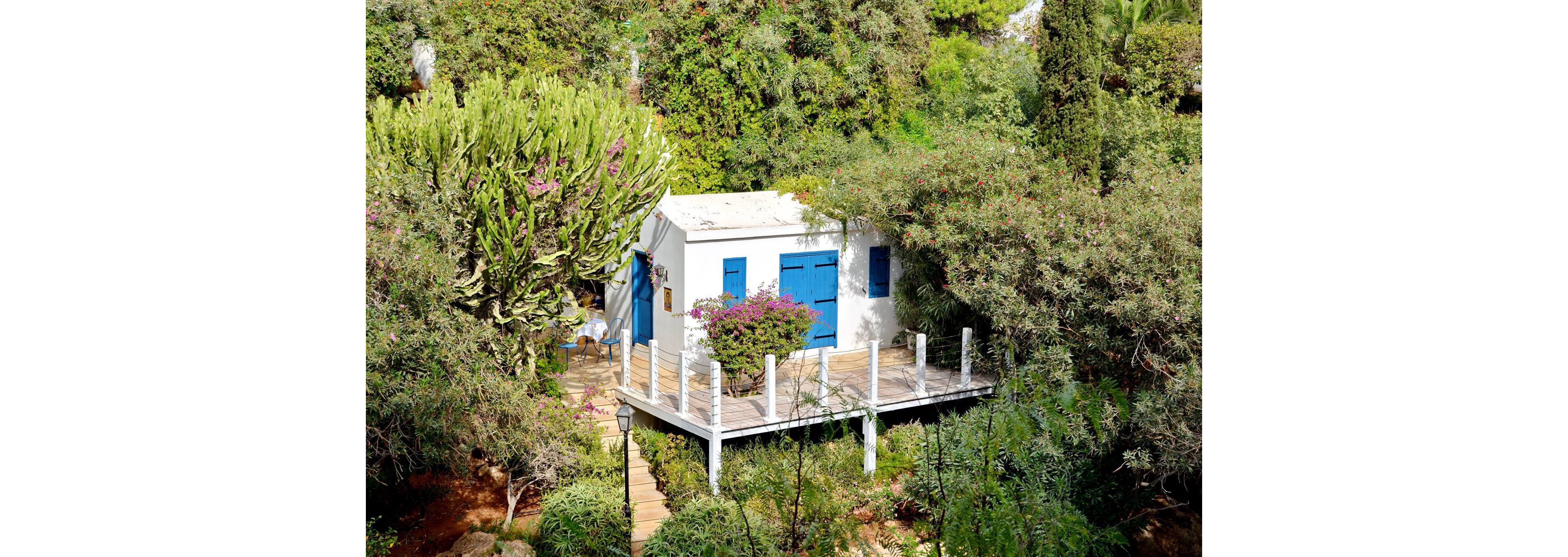 A small white cottage with bright blue doors and shutters sits elevated on stilts among dense greenery. A wooden deck and railing surround the front of the house, connected by a narrow pathway and steps leading through the garden. Lush trees, shrubs, and flowering plants frame the structure, creating a secluded, tranquil atmosphere. The scene suggests a peaceful retreat or vacation home nestled in nature.
