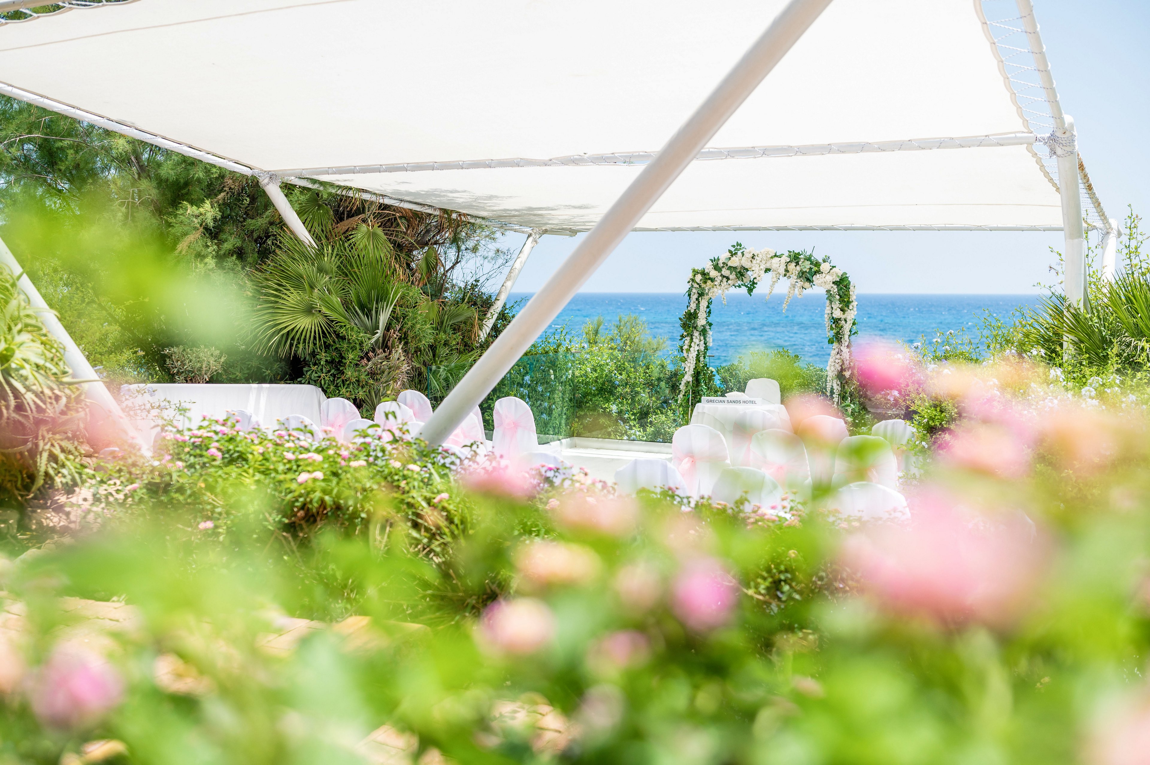 An elegant outdoor wedding ceremony setup is arranged beneath a white canopy overlooking the sea. A floral arch stands at the front, framed by lush greenery and palm trees. White chairs with soft pink accents are visible in the background, suggesting a romantic event. Blurred pink flowers in the foreground add depth and a dreamy, summery atmosphere.