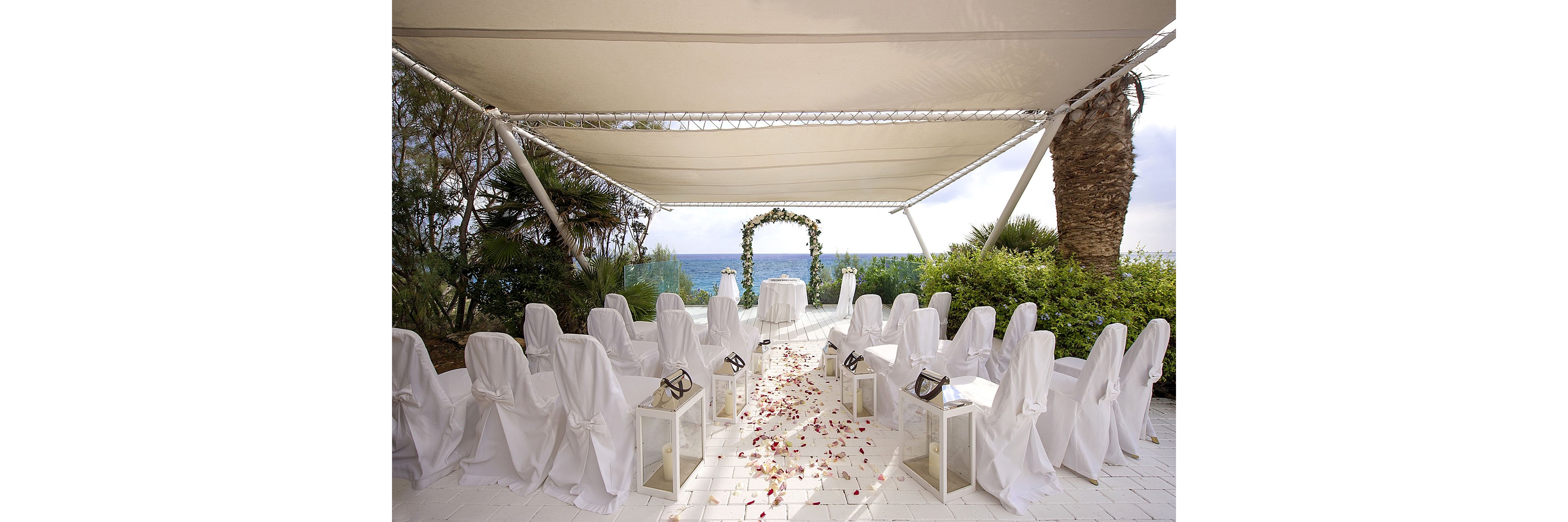 An elegant outdoor wedding ceremony setup is arranged under a white canopy overlooking the sea. Two rows of white chair covers face a floral arch framing the ocean horizon. Lanterns line the aisle, which is scattered with rose petals leading to a small table at the front. Lush greenery and palm trees surround the venue, creating a serene coastal atmosphere.