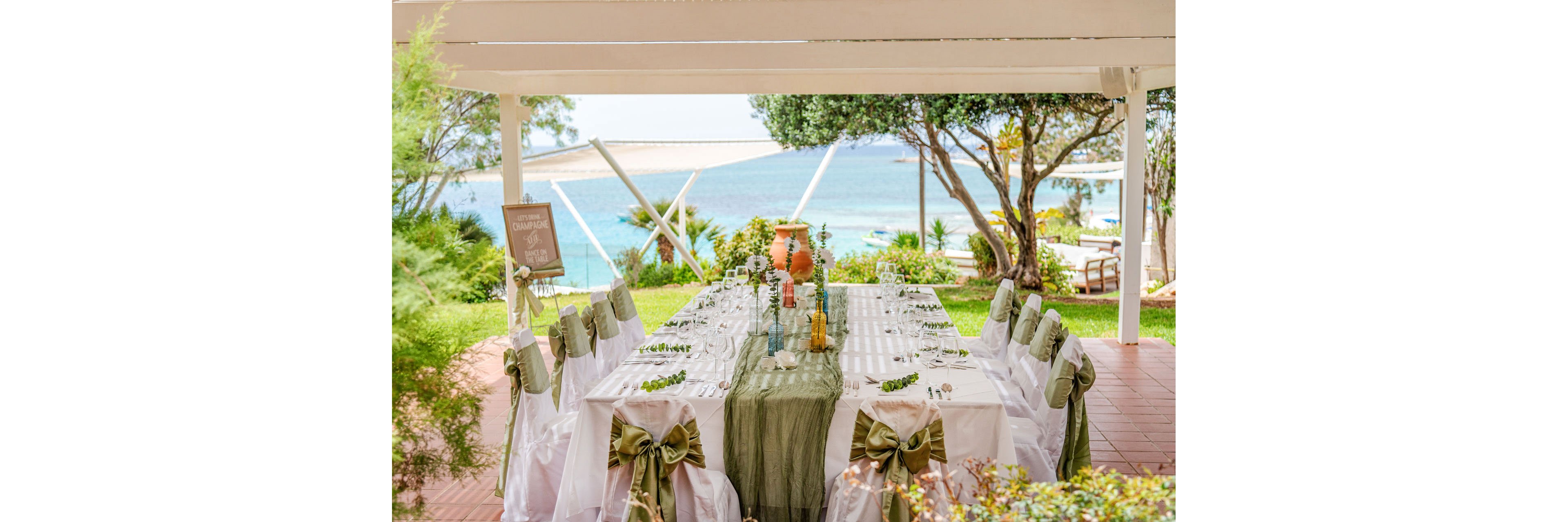 A long banquet table is beautifully set for a formal event under a white pergola by the sea. White chairs with green satin bows line both sides of the table, which is decorated with a green table runner and glassware. Floral arrangements and rustic vases create a romantic centerpiece, while greenery surrounds the terrace. In the background, blue ocean water and sunlit sky enhance the serene coastal atmosphere.